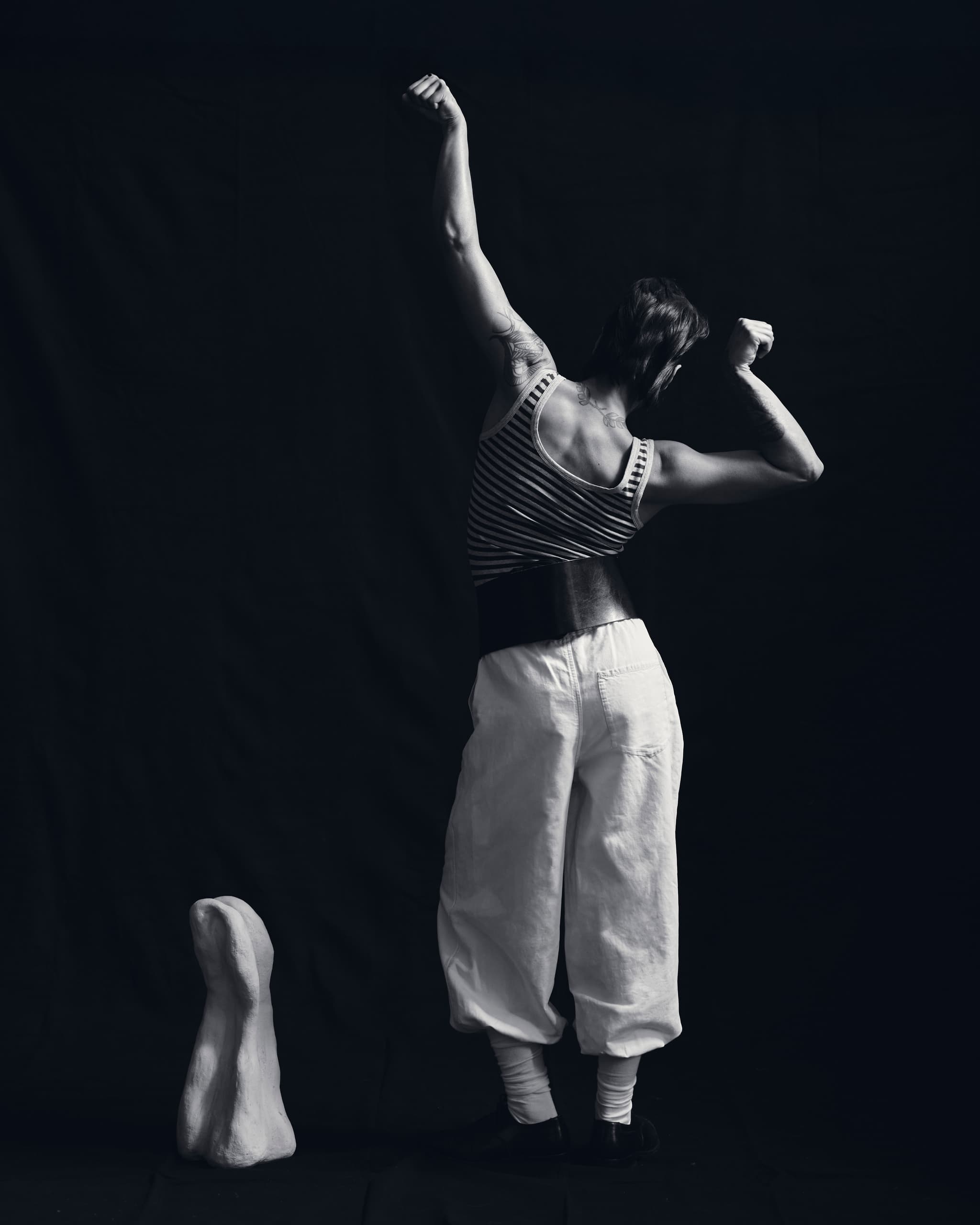 Person flexing muscles in striped top against black background, next to a sculpture.