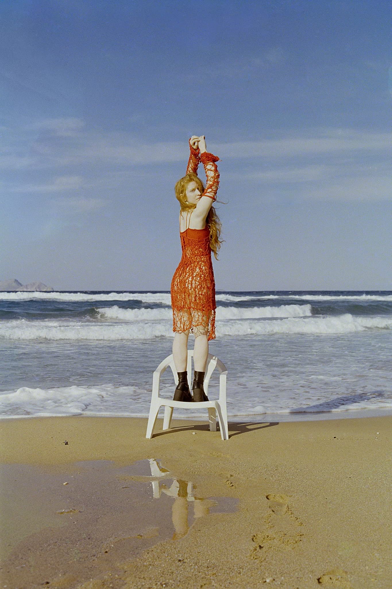 Person in red dress on chair at beach, arms raised, waves and blue sky in background.