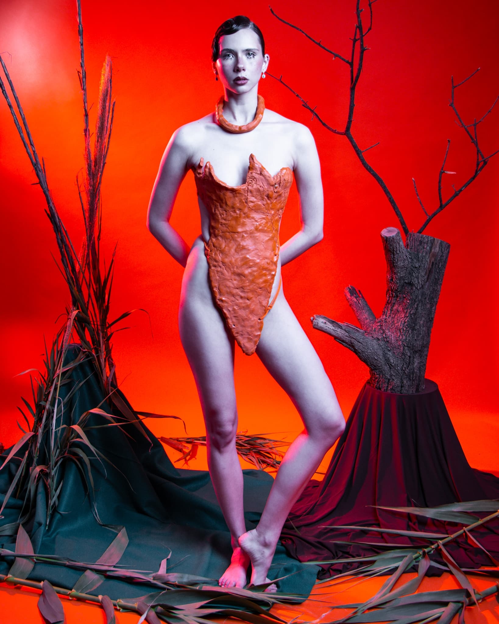 Model in avant-garde orange outfit with red backdrop and nature elements.