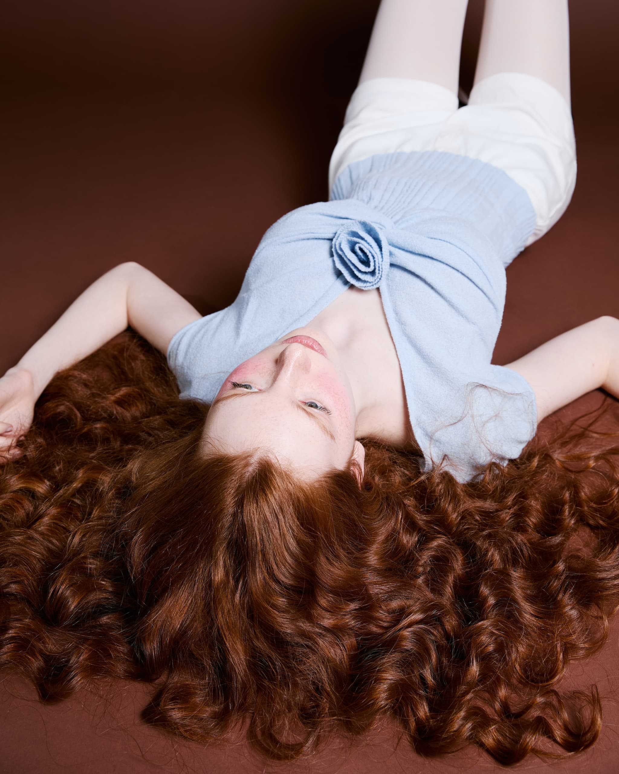 Person with long, curly, red hair lying on a brown surface, wearing a blue top and shorts.