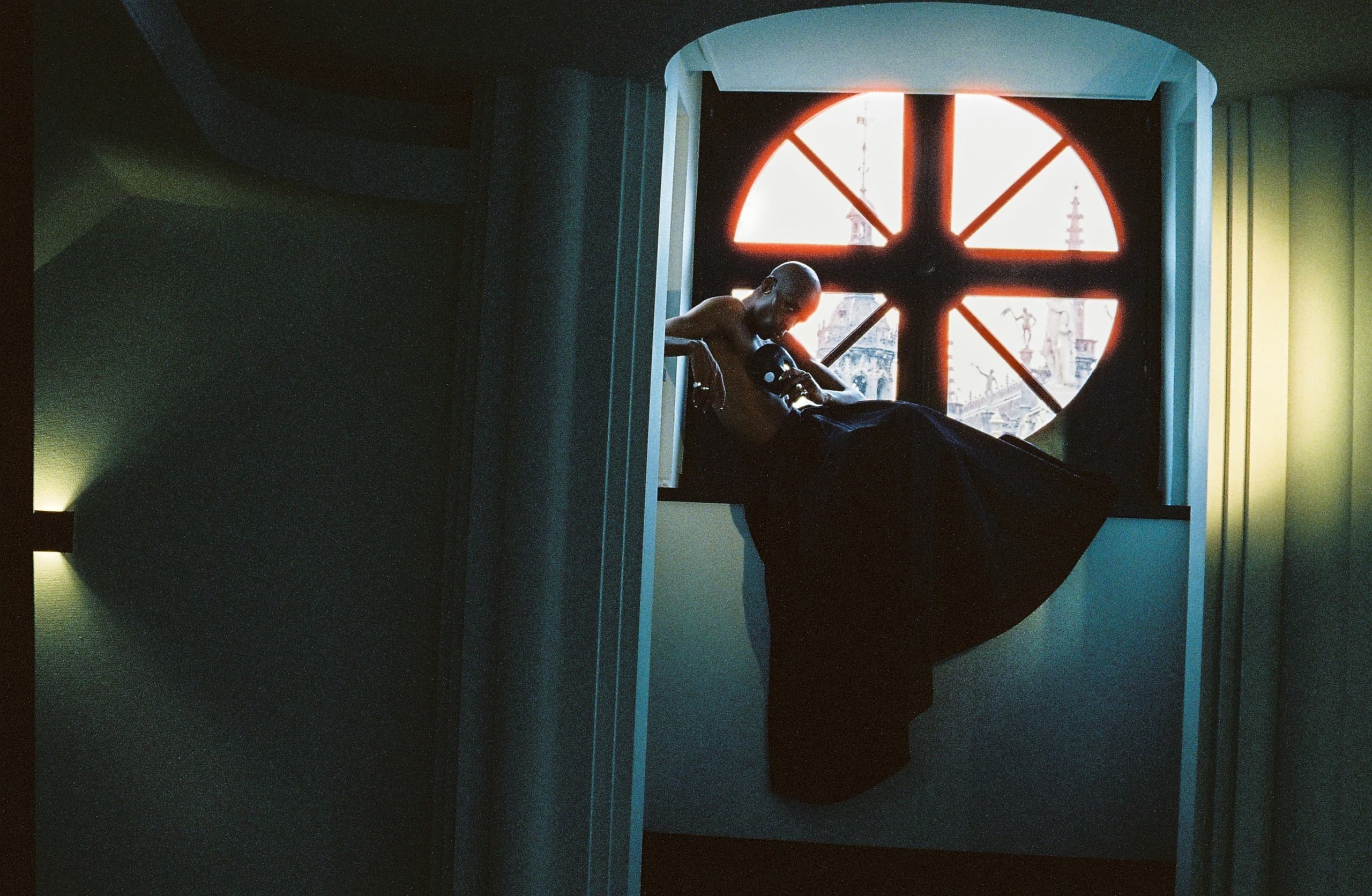 Person silhouetted by round window, sitting with violin, in moody dim light.