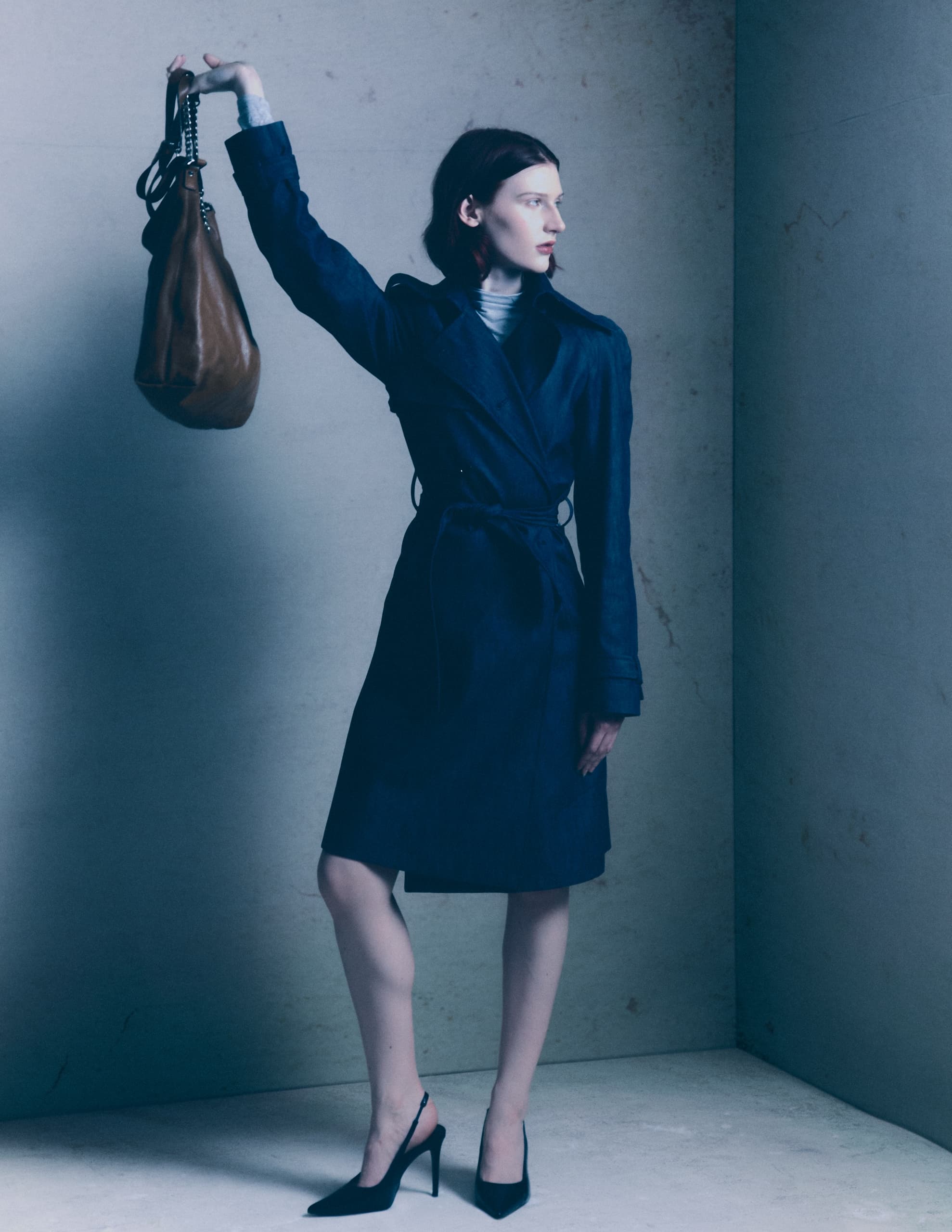 Fashion model in trench coat holding a stylish handbag indoors.