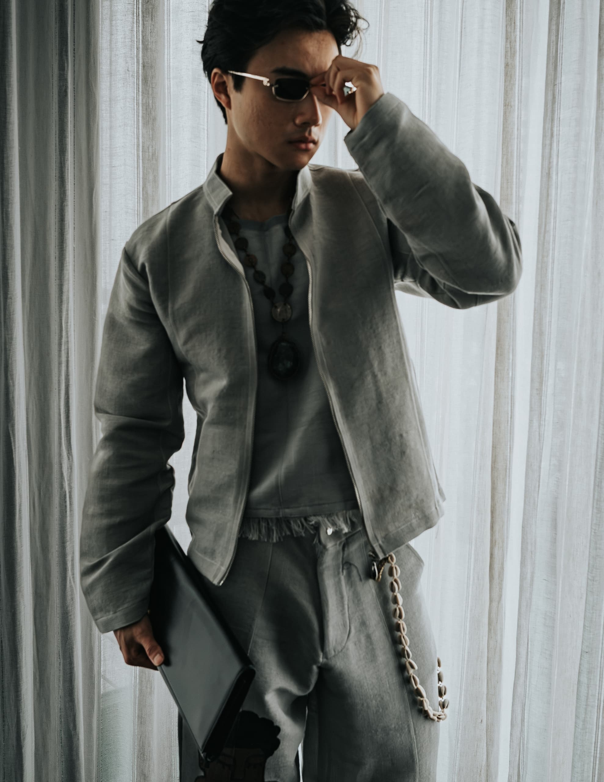 Stylish man in gray attire with sunglasses and laptop, posing by sheer curtains.