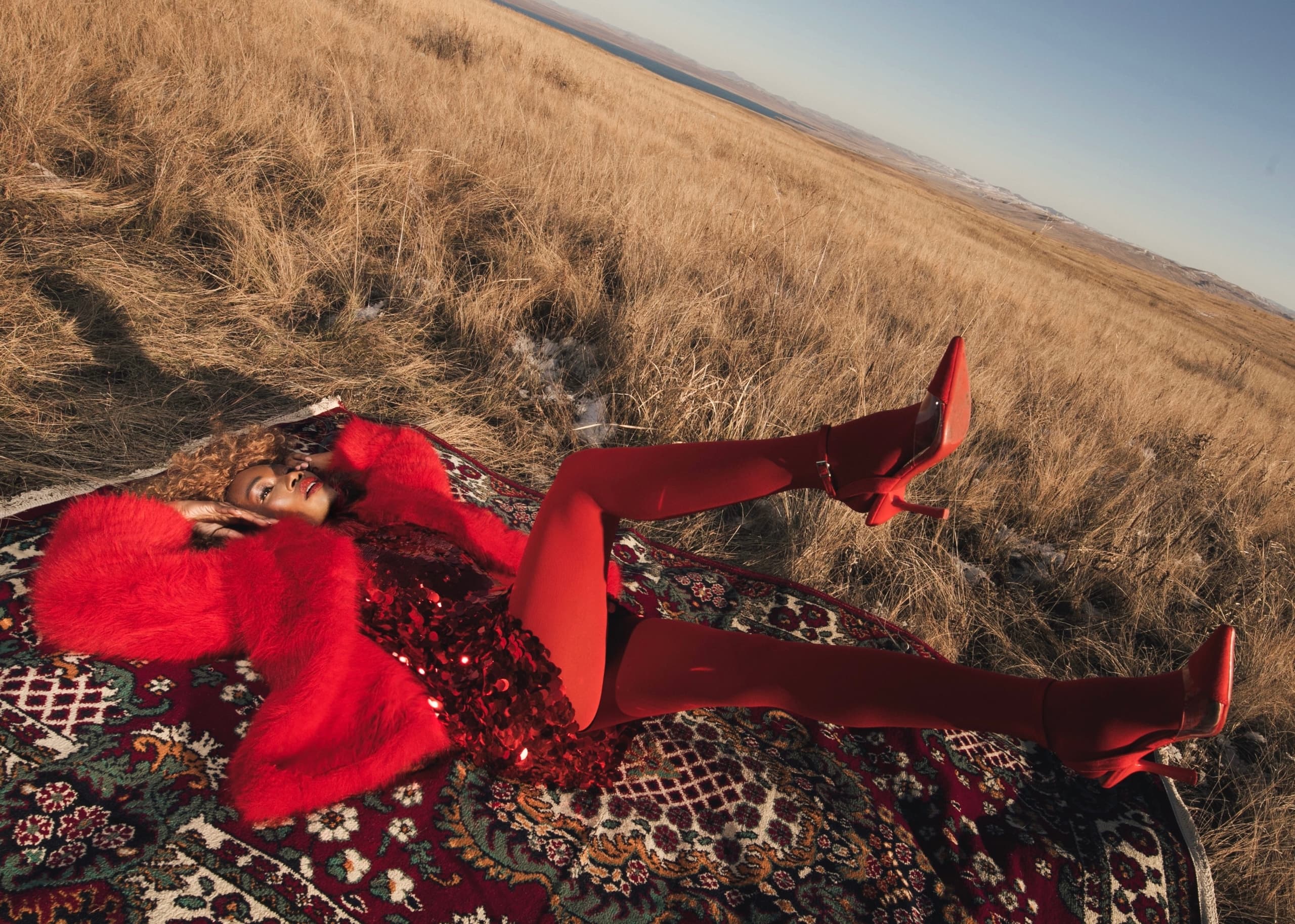 Woman in red fashion outfit lying on a patterned rug in a vast grassy field.