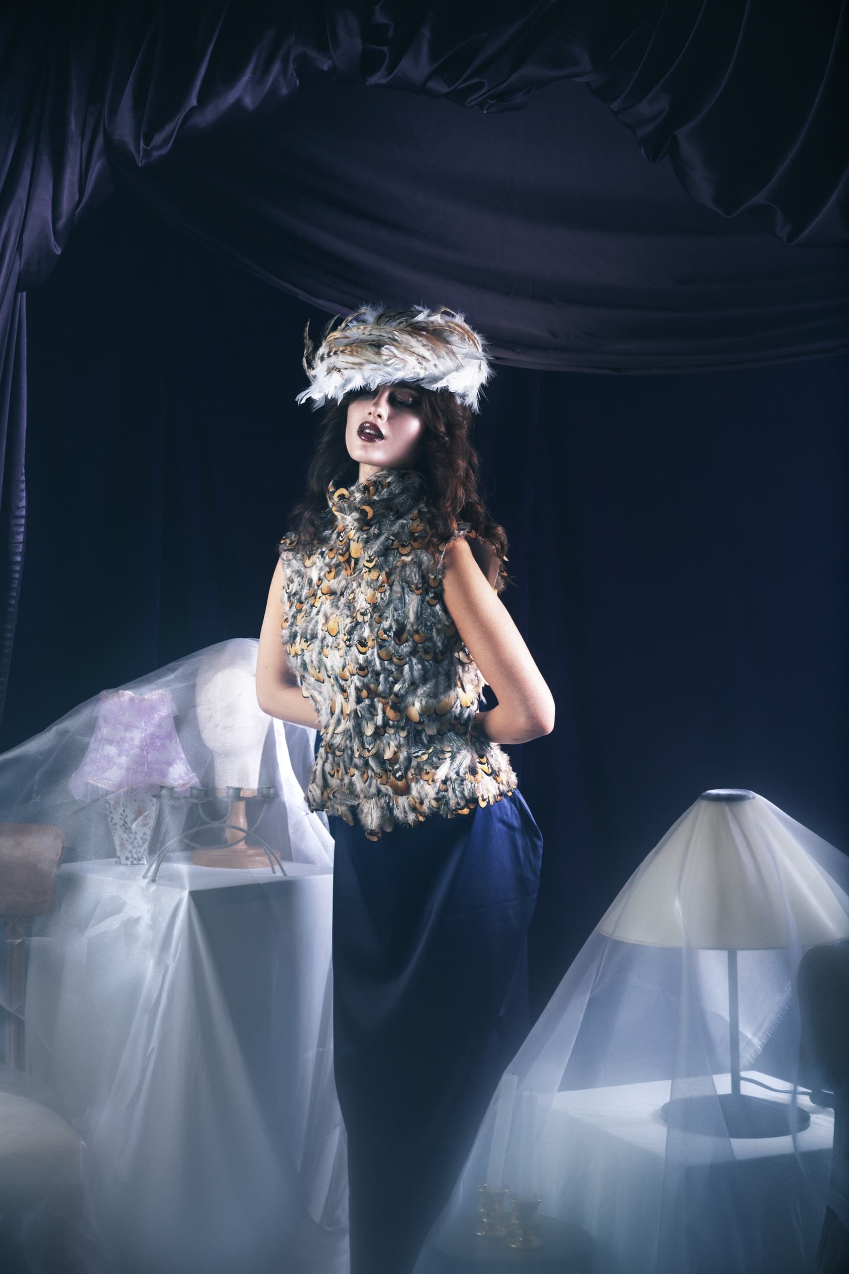 Elegant model in feather outfit and hat, surrounded by draped fabric and soft lighting.