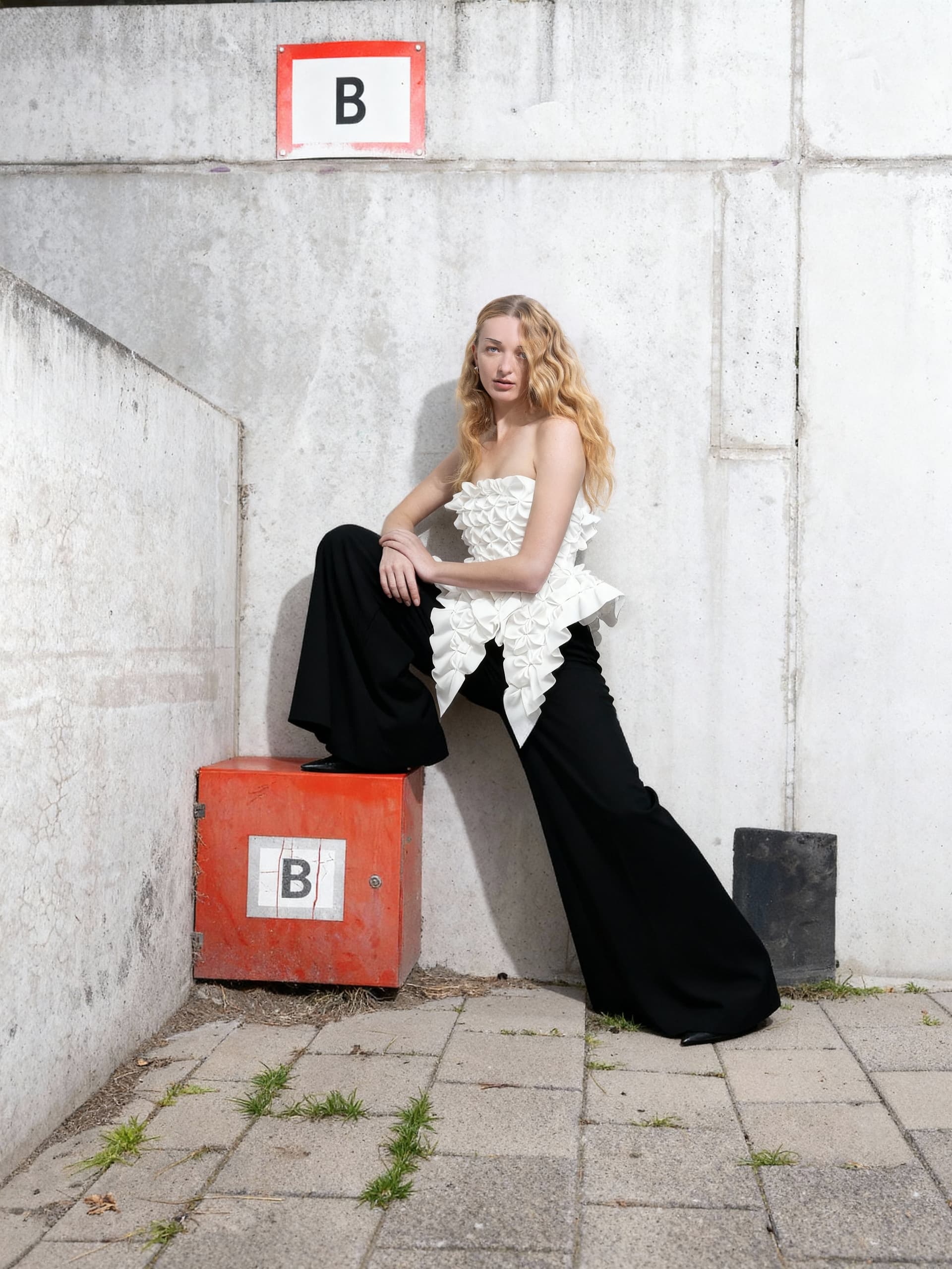 Fashionable woman in white top and black pants posing against urban concrete backdrop.