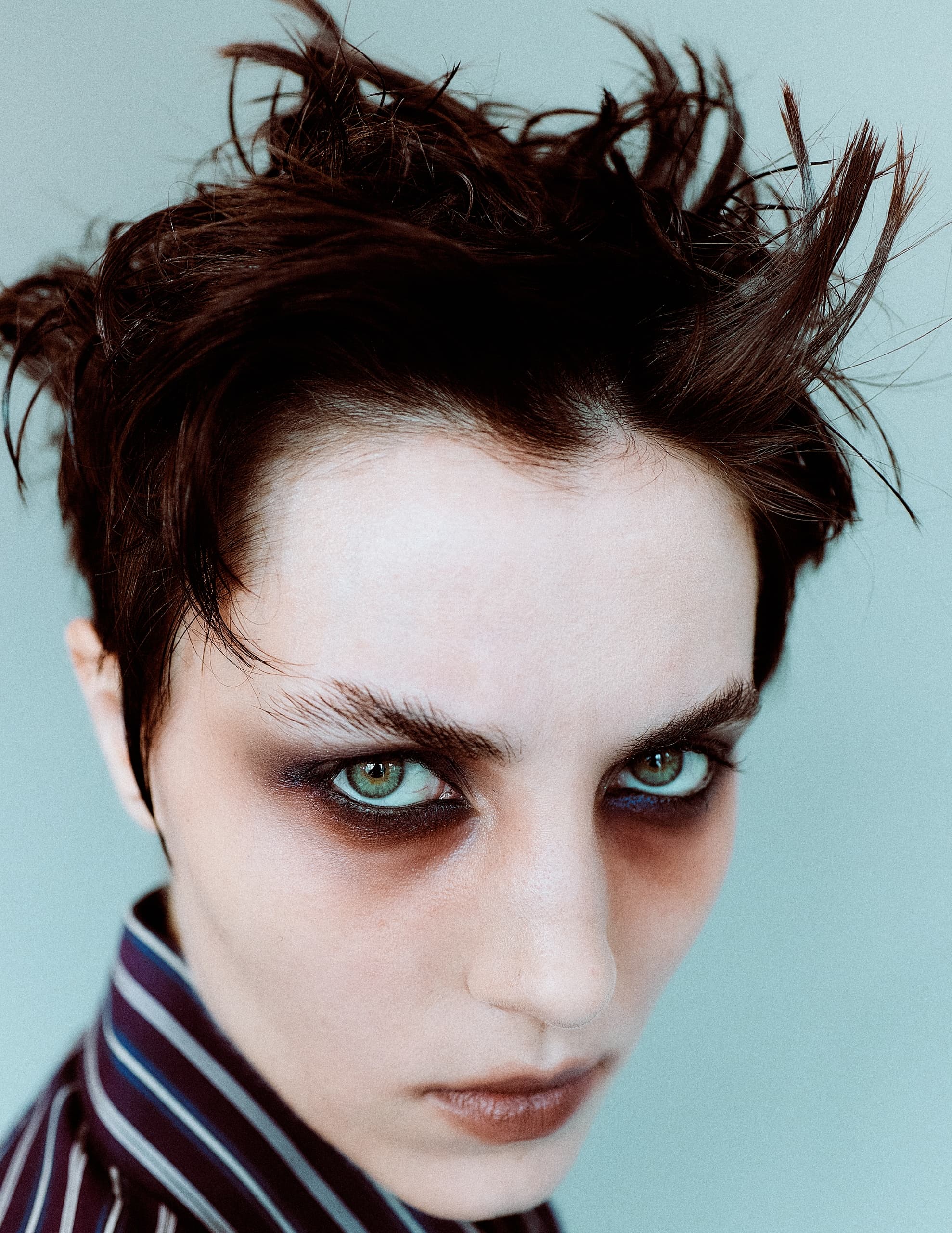 Edgy portrait of person with dramatic eye makeup and spiky hair, wearing striped shirt.
