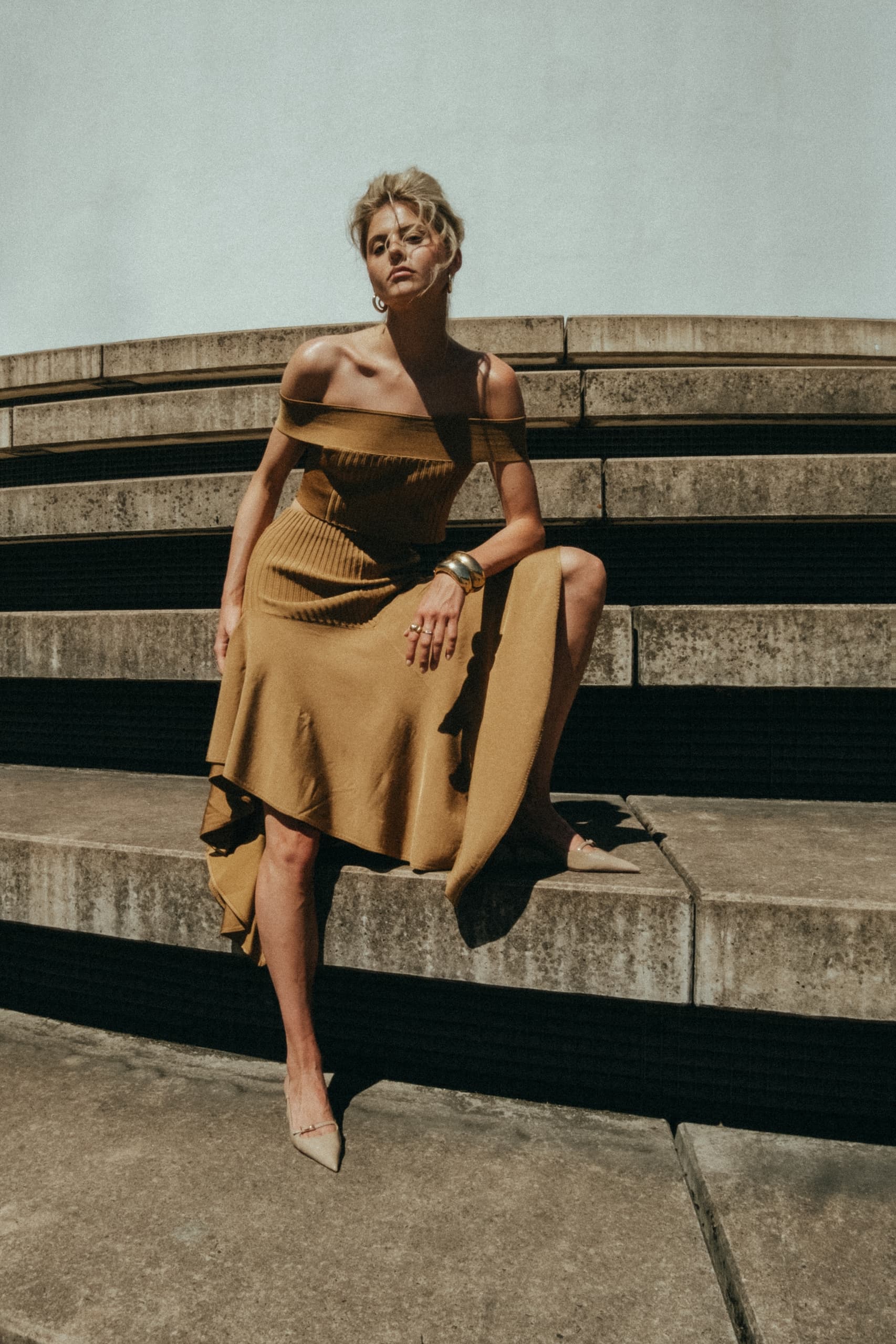 Woman in mustard dress sitting on concrete stairs, sunlit urban fashion.