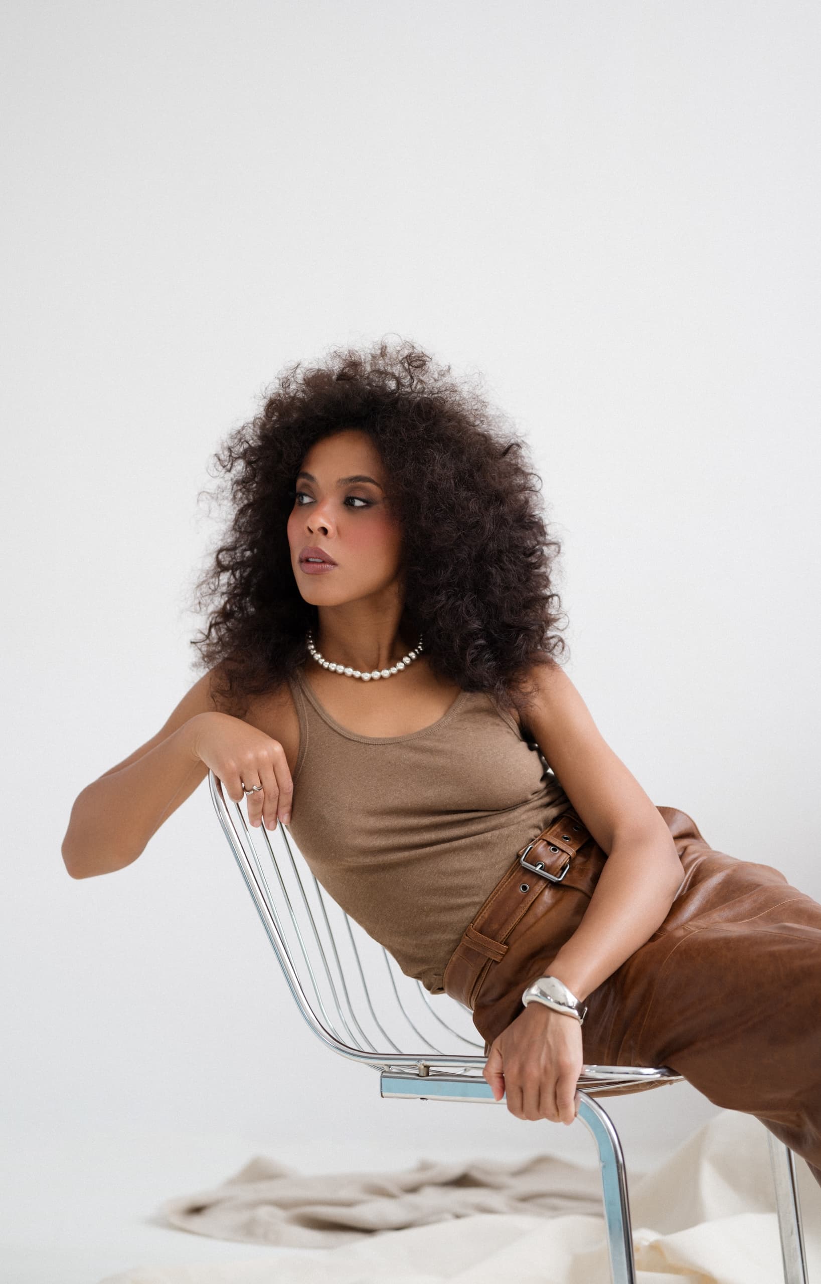 Woman with curly hair in brown outfit, posing on a chair with stylish jewelry.