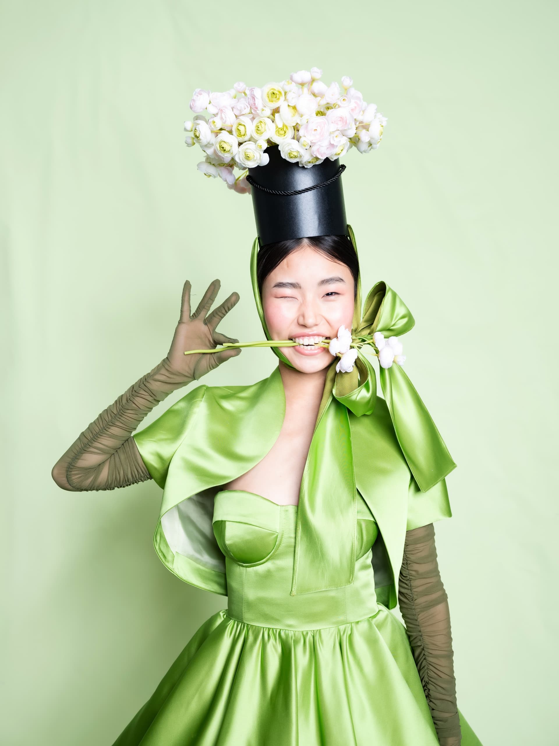Woman in green dress and floral hat winks while posing with flower in mouth.