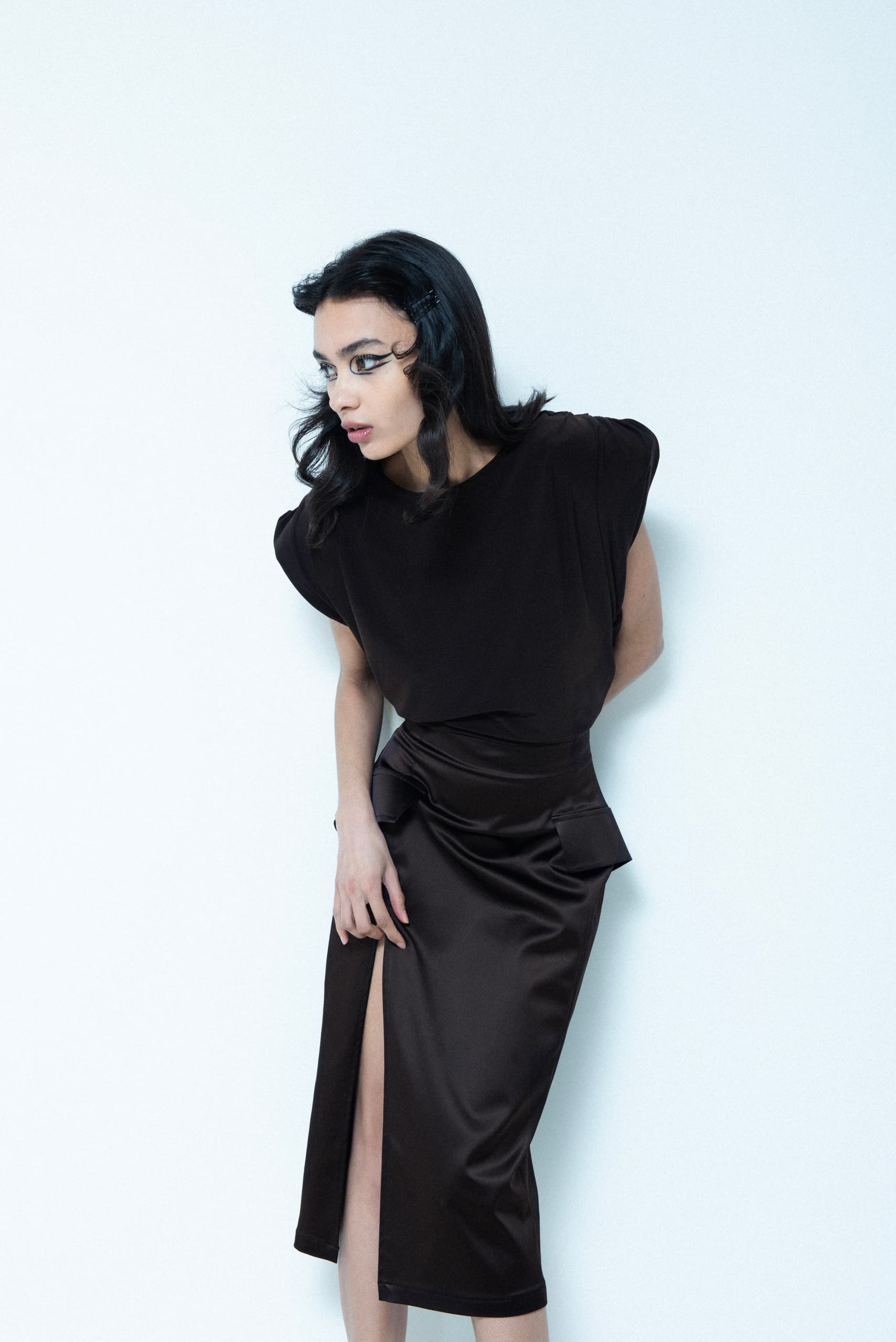 Stylish woman in black dress with slit, striking fashion pose against white backdrop.