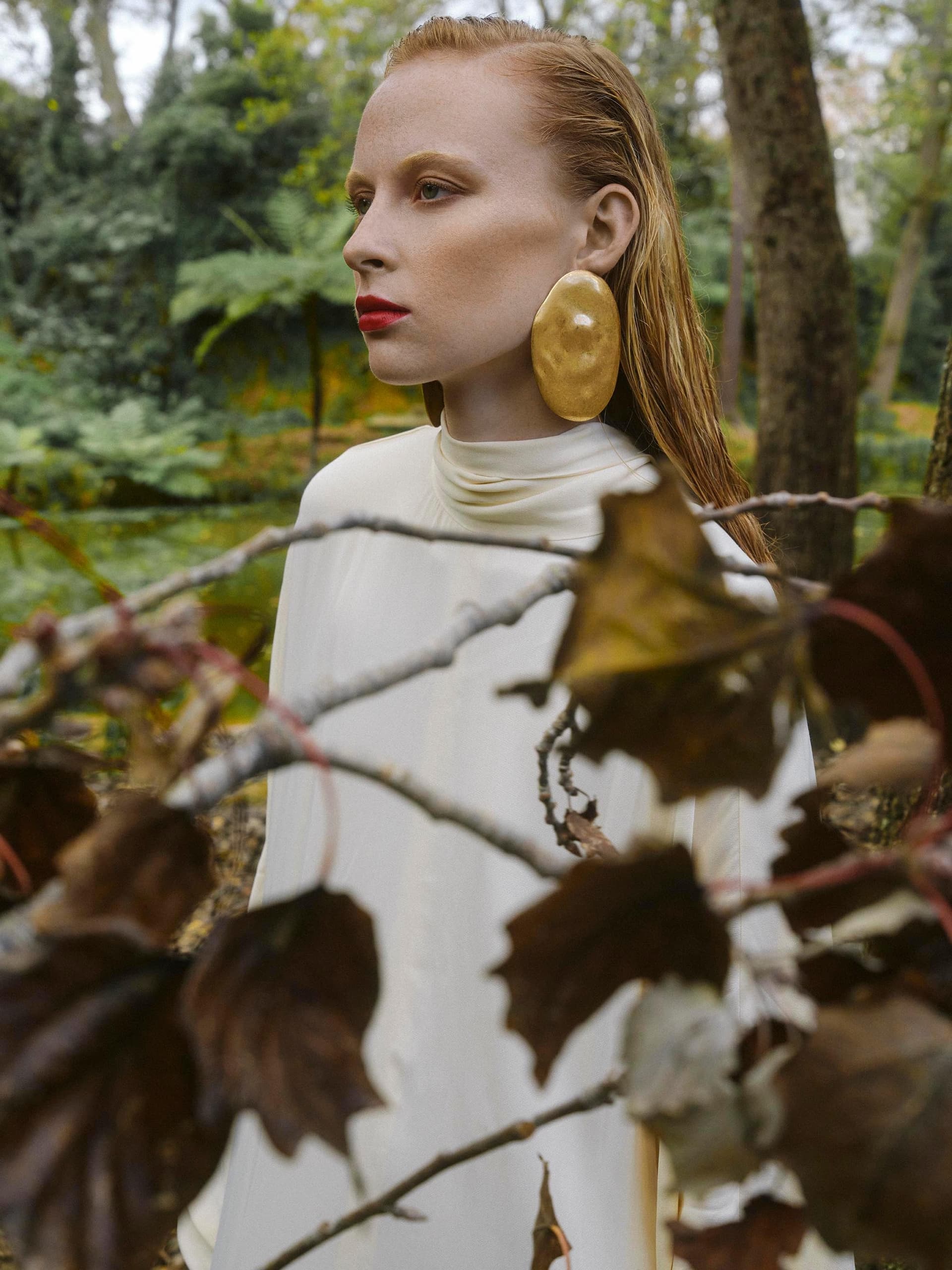 Woman in forest with bold earrings and red lipstick, wearing a white dress.
