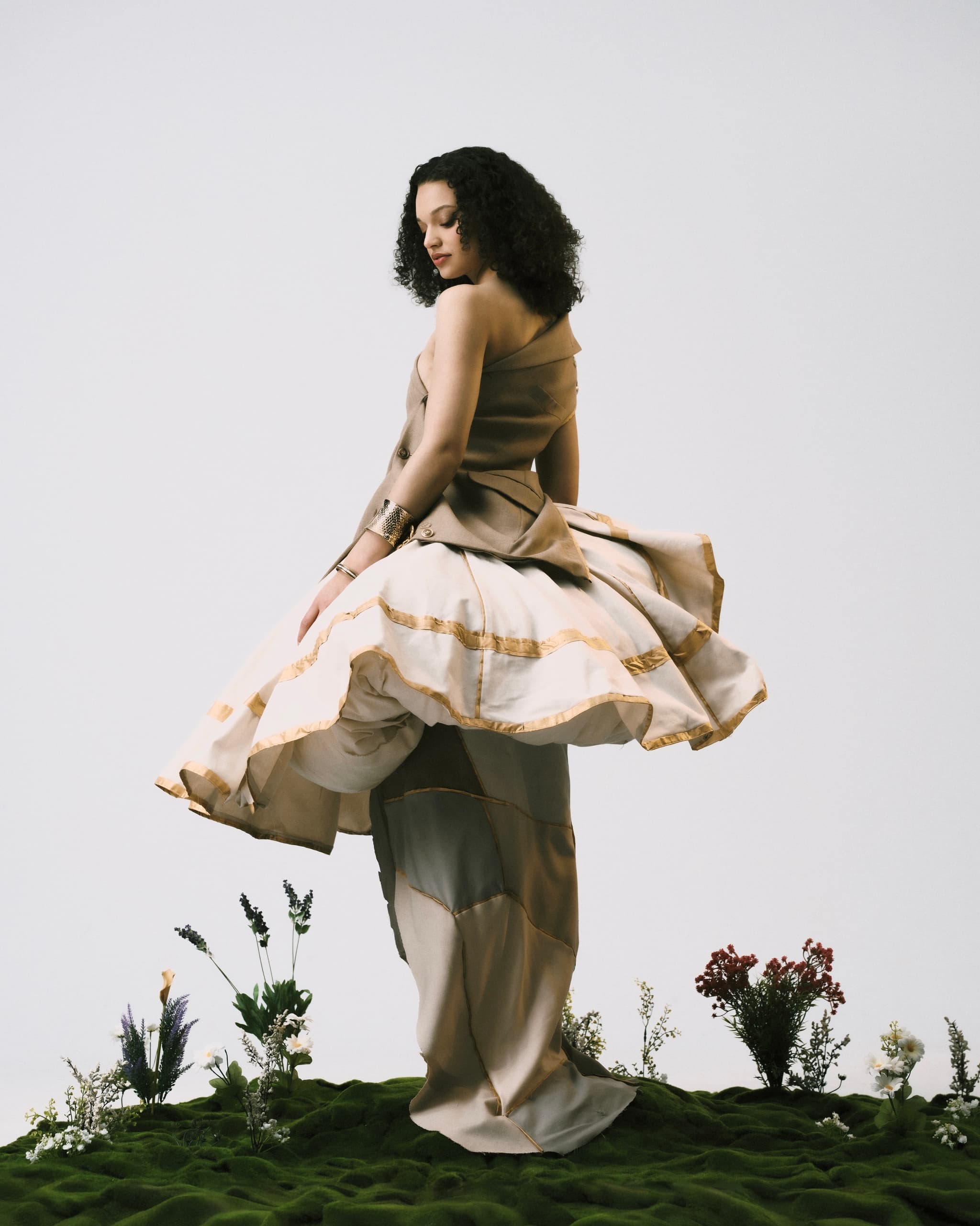 Elegant woman in flowing dress stands on moss, surrounded by flowers.