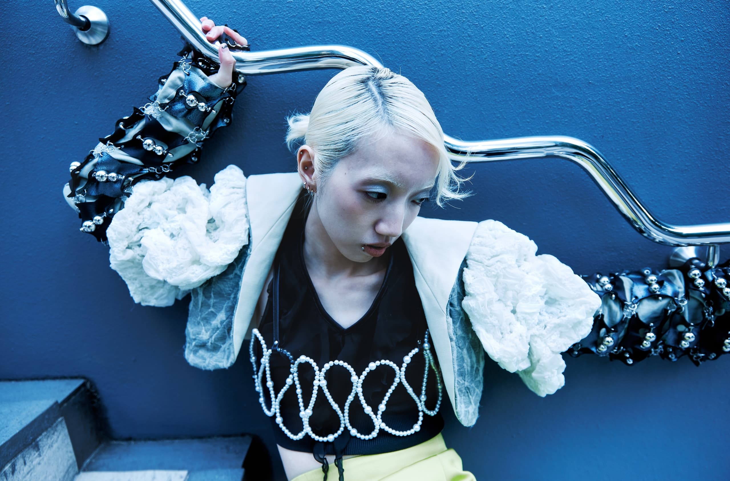 Fashionable person in avant-garde outfit poses against a blue wall with metal railing.