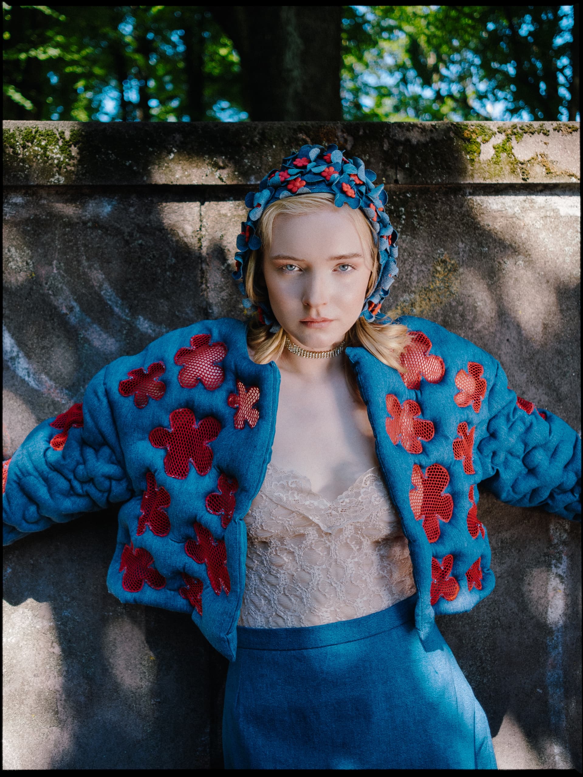 Woman in blue floral jacket and bonnet standing against a stone wall.