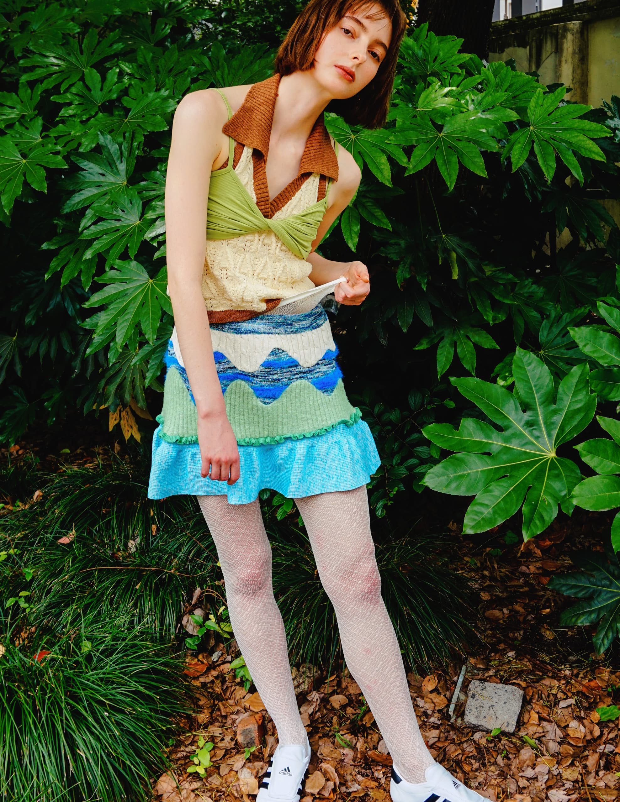 Woman in eclectic fashion poses in leafy outdoor setting.