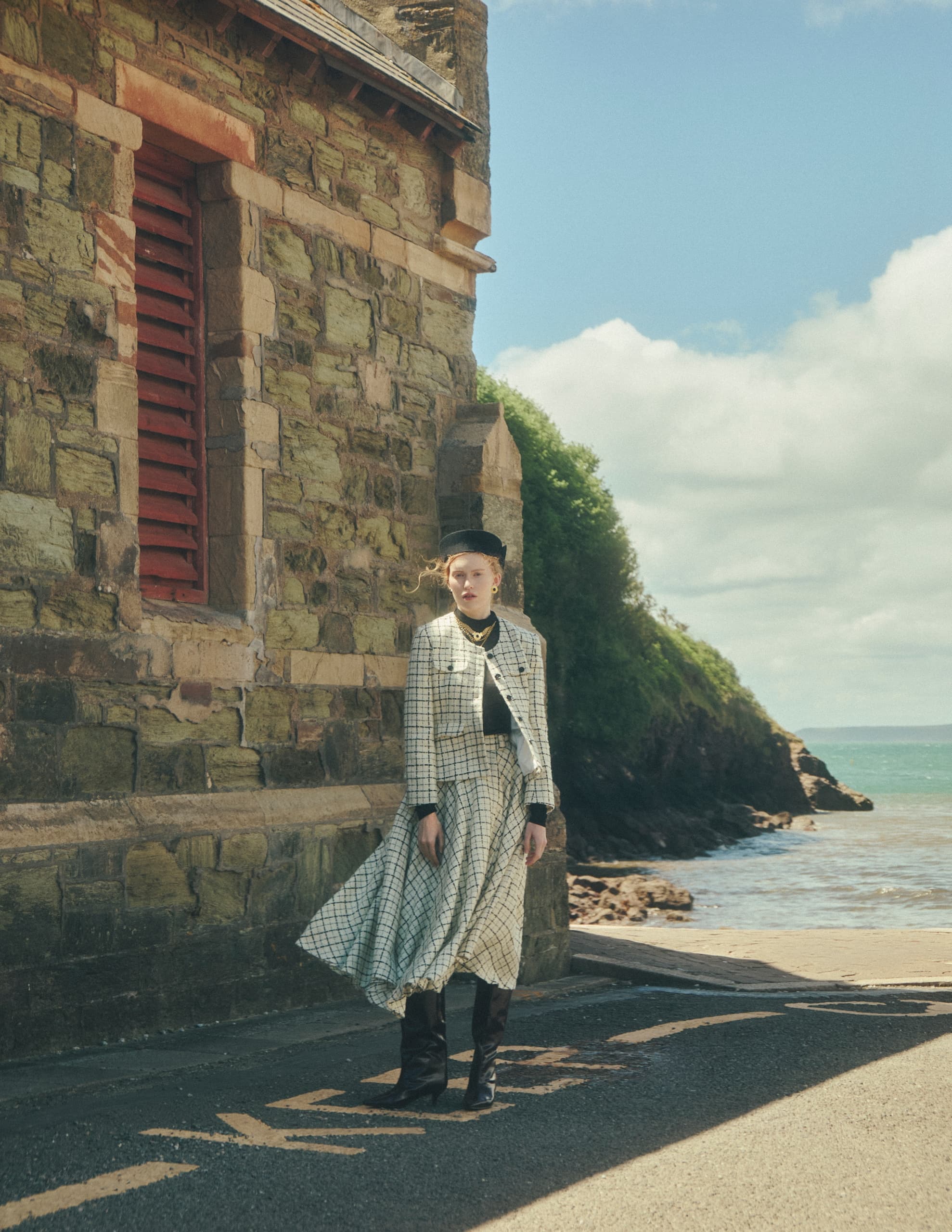 Woman in vintage attire stands by a seaside stone building on a sunny day.