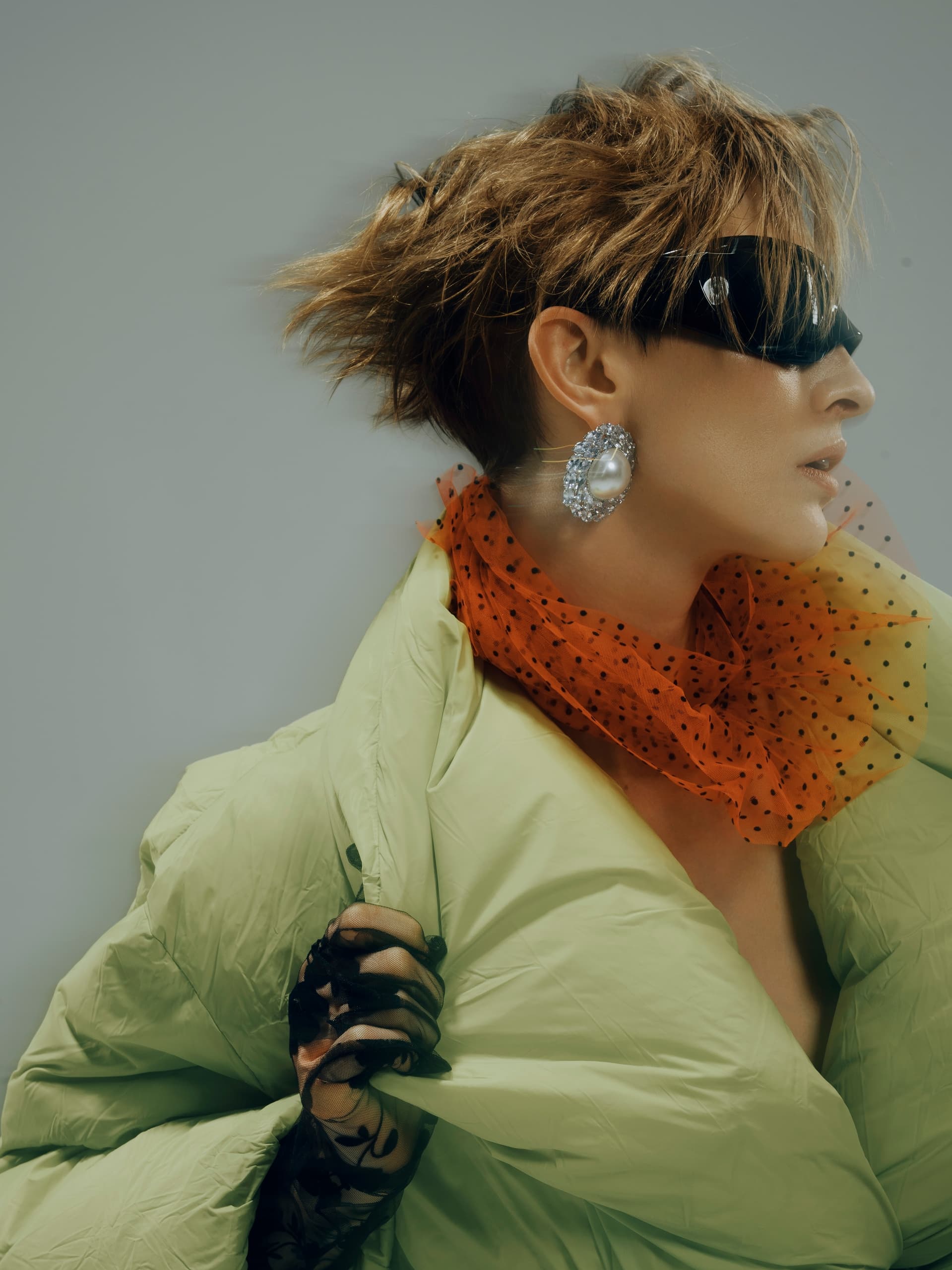 Fashionable woman in green coat, orange scarf, and bold sunglasses and earrings.