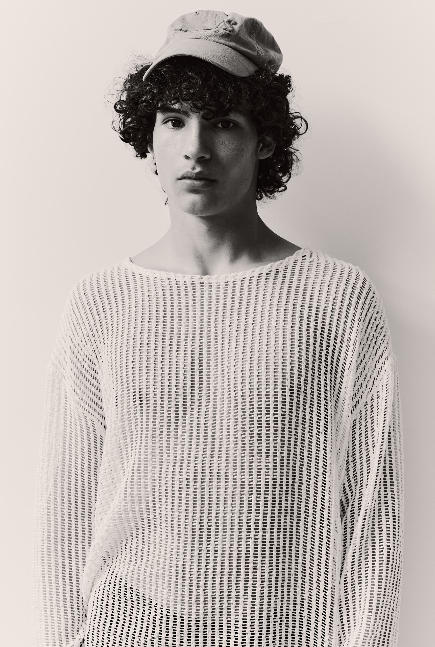 Young person with curly hair wearing a mesh sweater and cap, black and white portrait.
