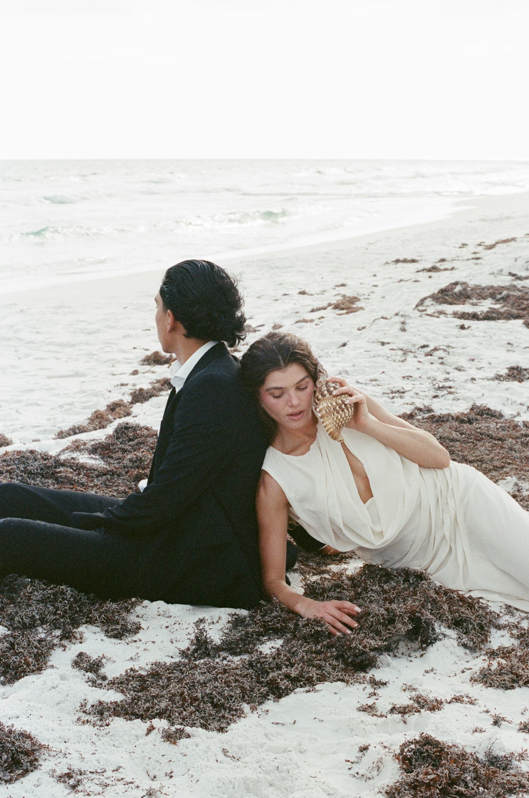 Couple in formal attire sitting on a seaweed-covered beach by the ocean.