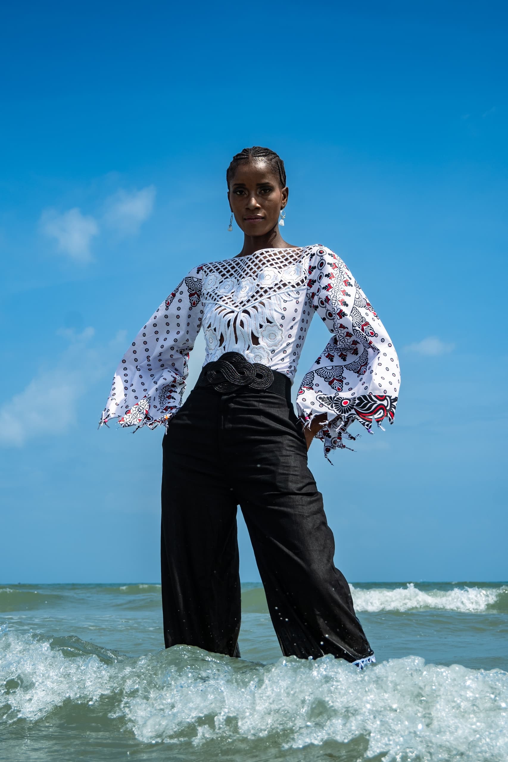 Model in stylish outfit standing in ocean waves under a clear blue sky.