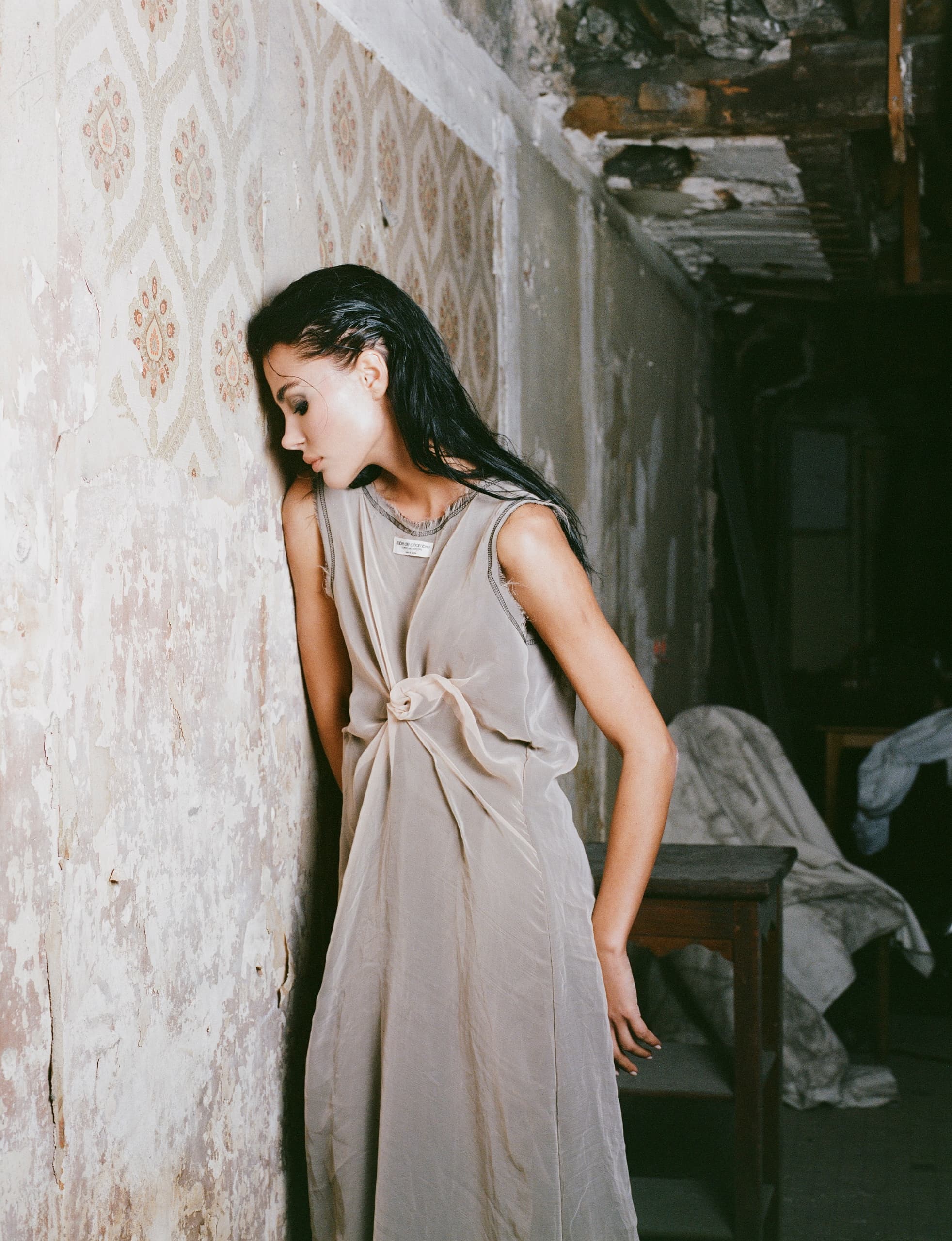 Woman in elegant gown leaning against vintage wall in dimly lit room.