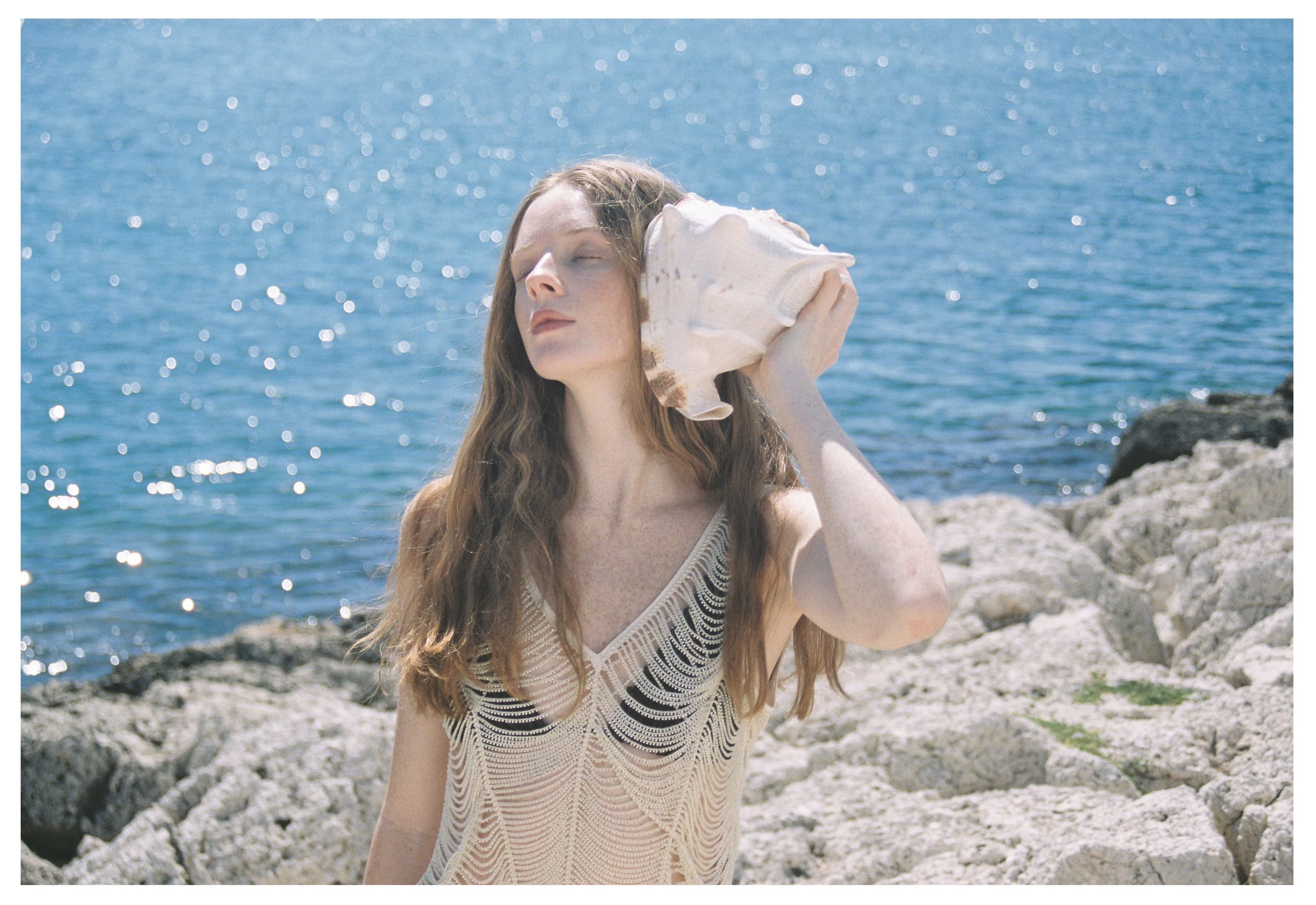 Woman enjoying ocean breeze with seashell on rocky shore.