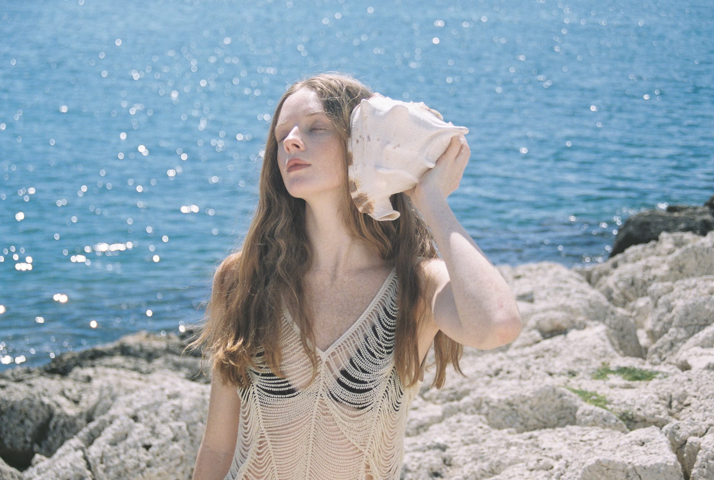 Woman by the sea holding a conch shell, embraced by sun and ocean breeze.