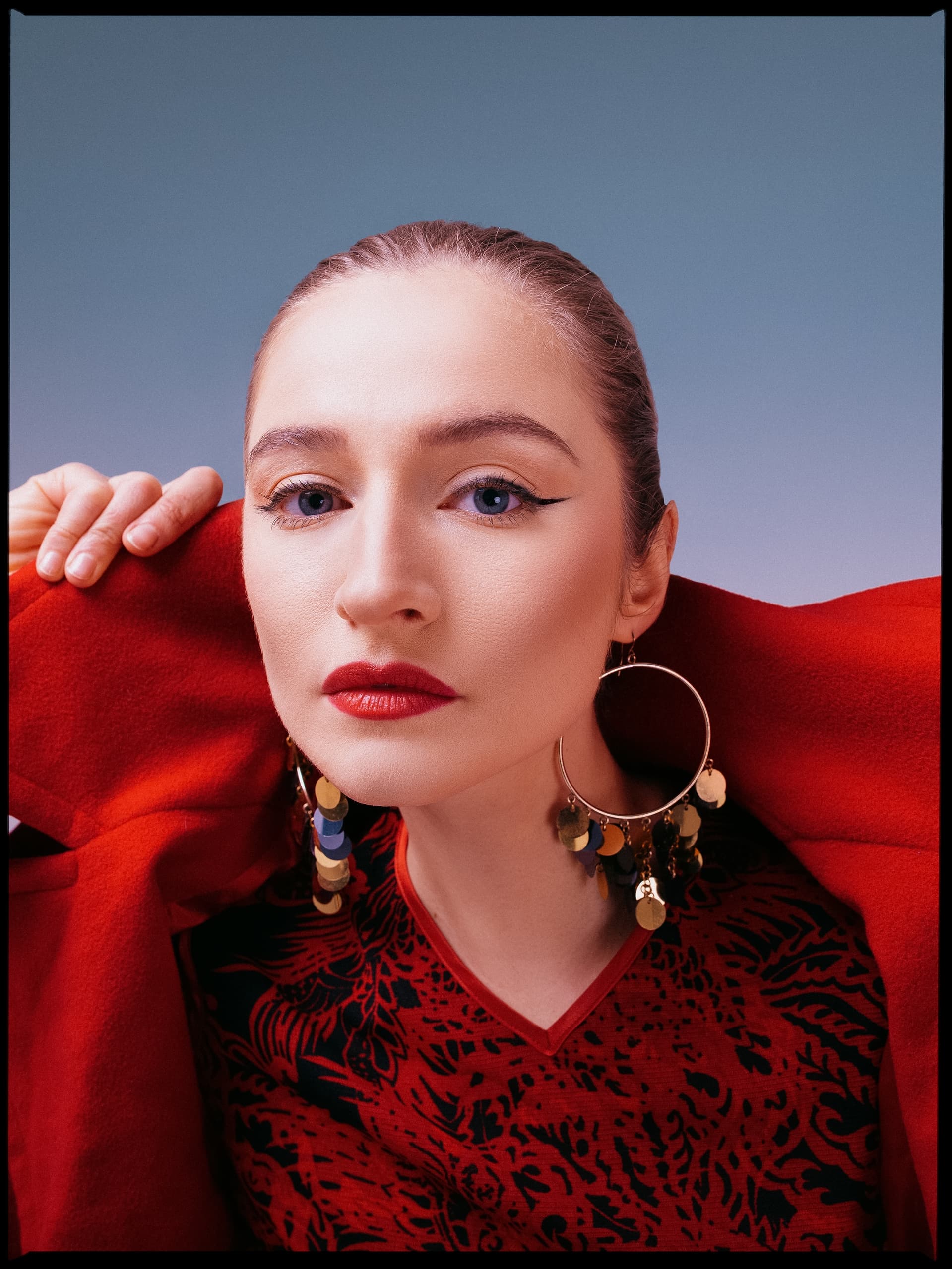 Woman in red coat and bold earrings with striking makeup against a blue background.
