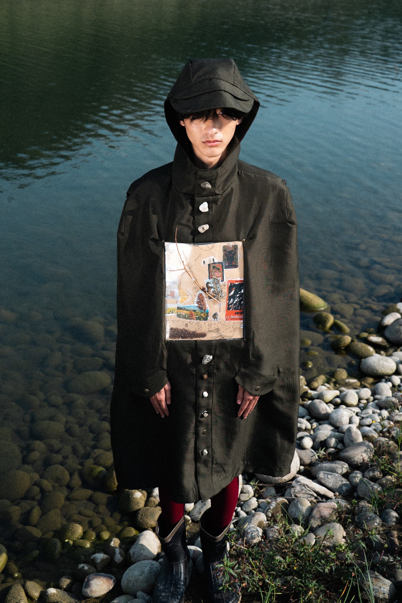 Person in artistic coat and hood standing by a rocky riverside.