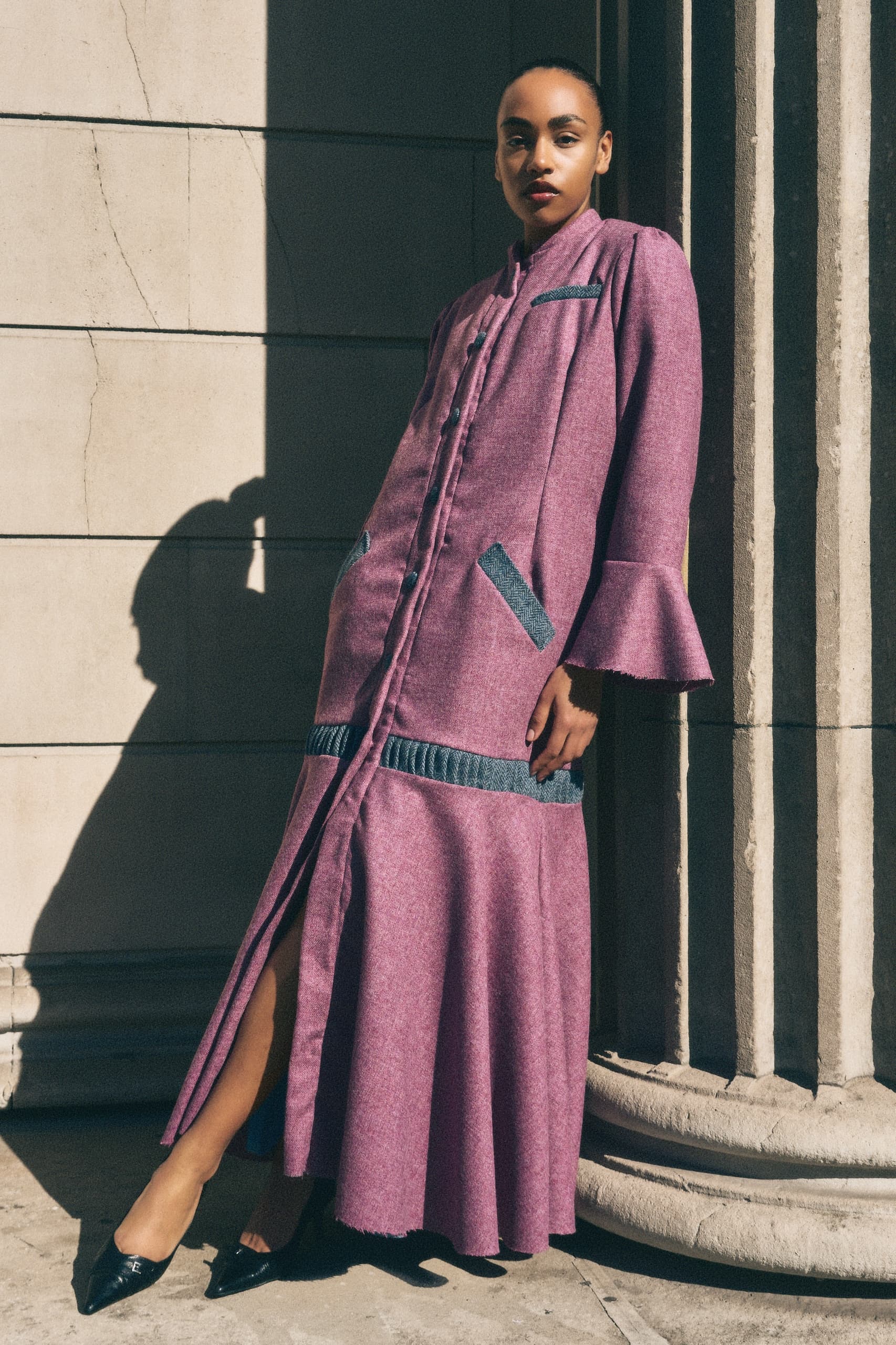 Woman in purple coat poses in sunlight against stone columns.
