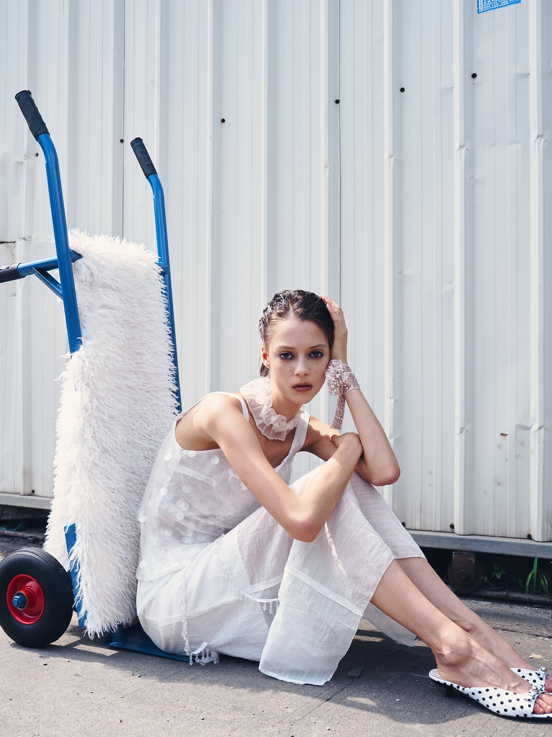 Model in white dress sitting outdoors, leaning against a fluffy cart.