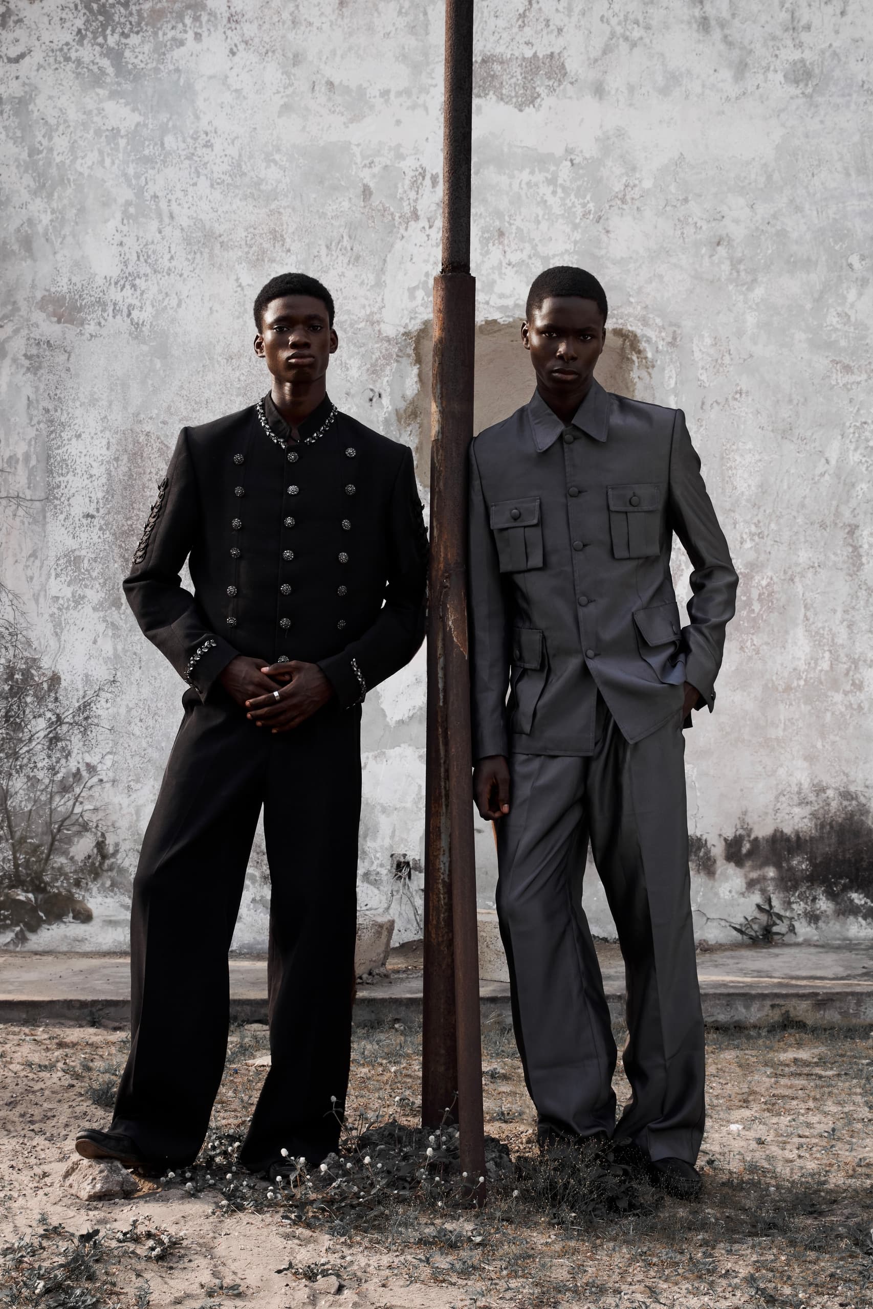 Two men in stylish black and gray suits pose against a textured wall outdoors.