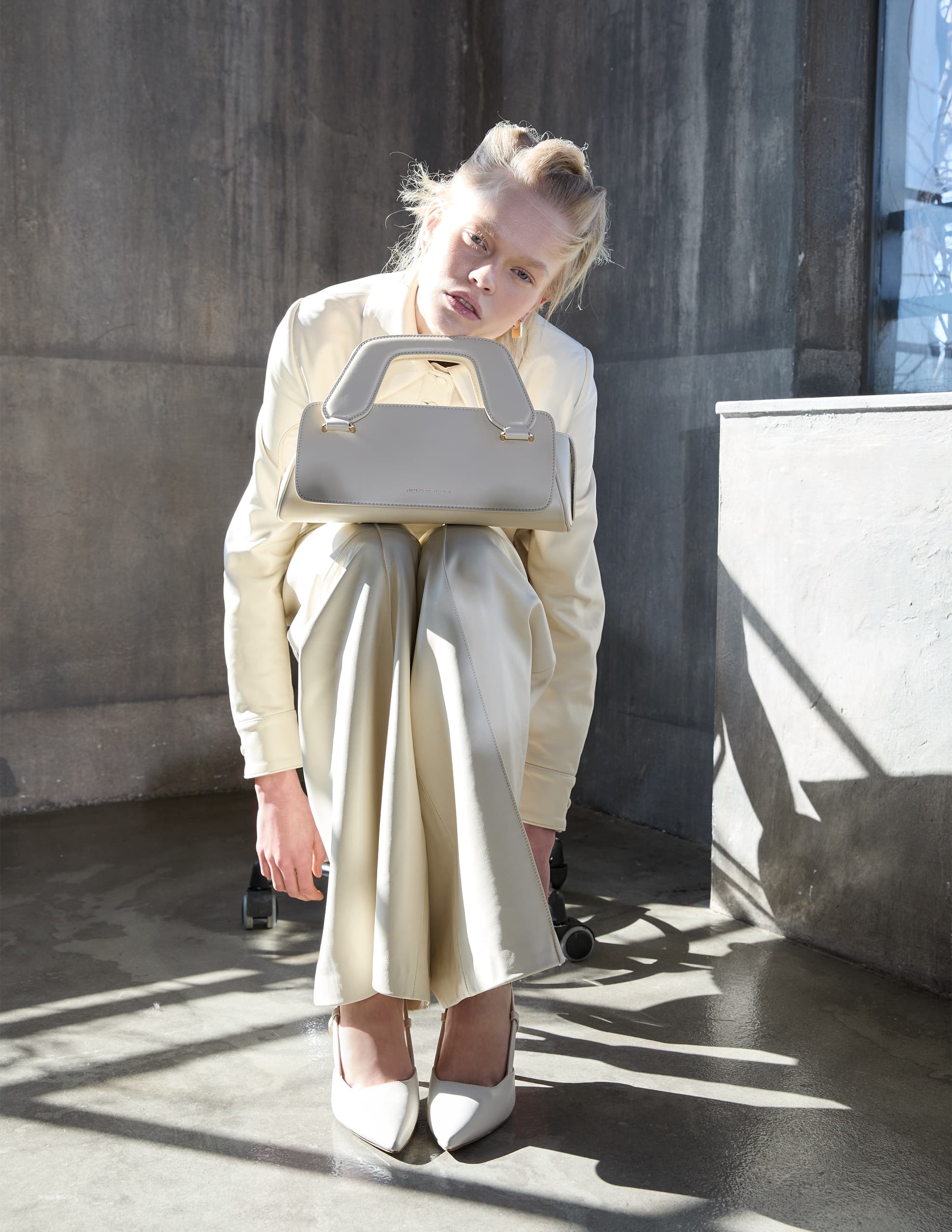 Fashion model holding beige handbag, dressed in cream outfit in bright sunlight.