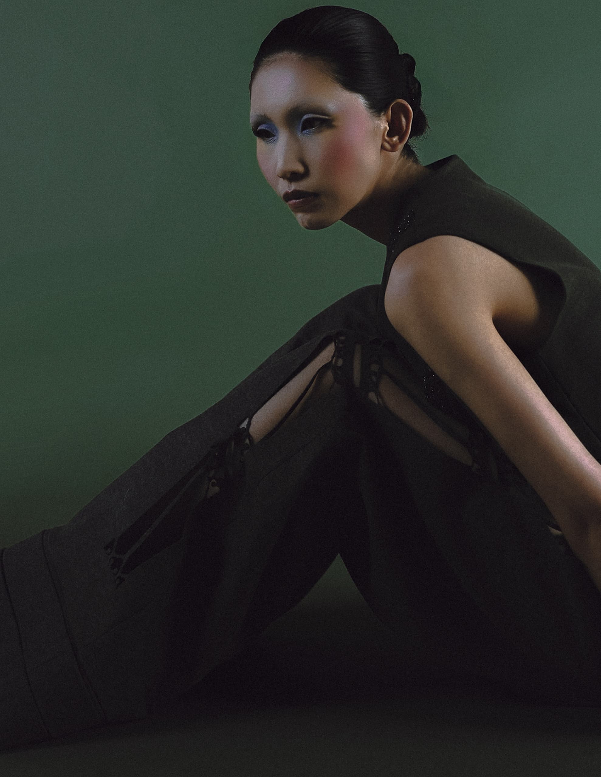 Fashion model in dark outfit with artistic makeup on a green background.