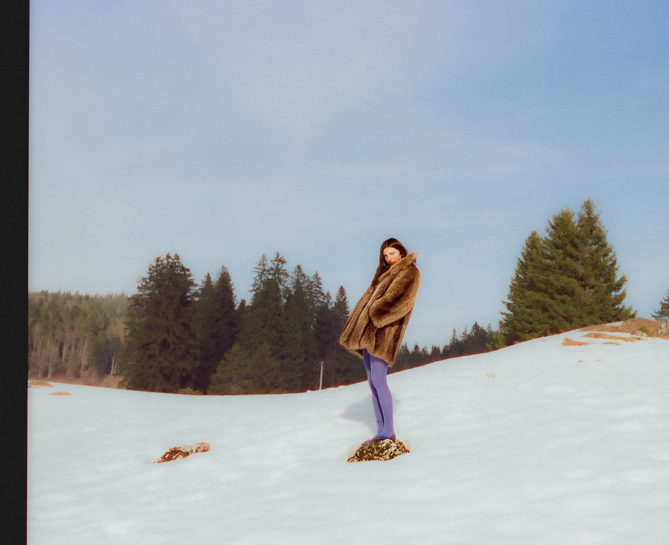 Woman in fur coat standing on snowy hill with pine trees in the background.