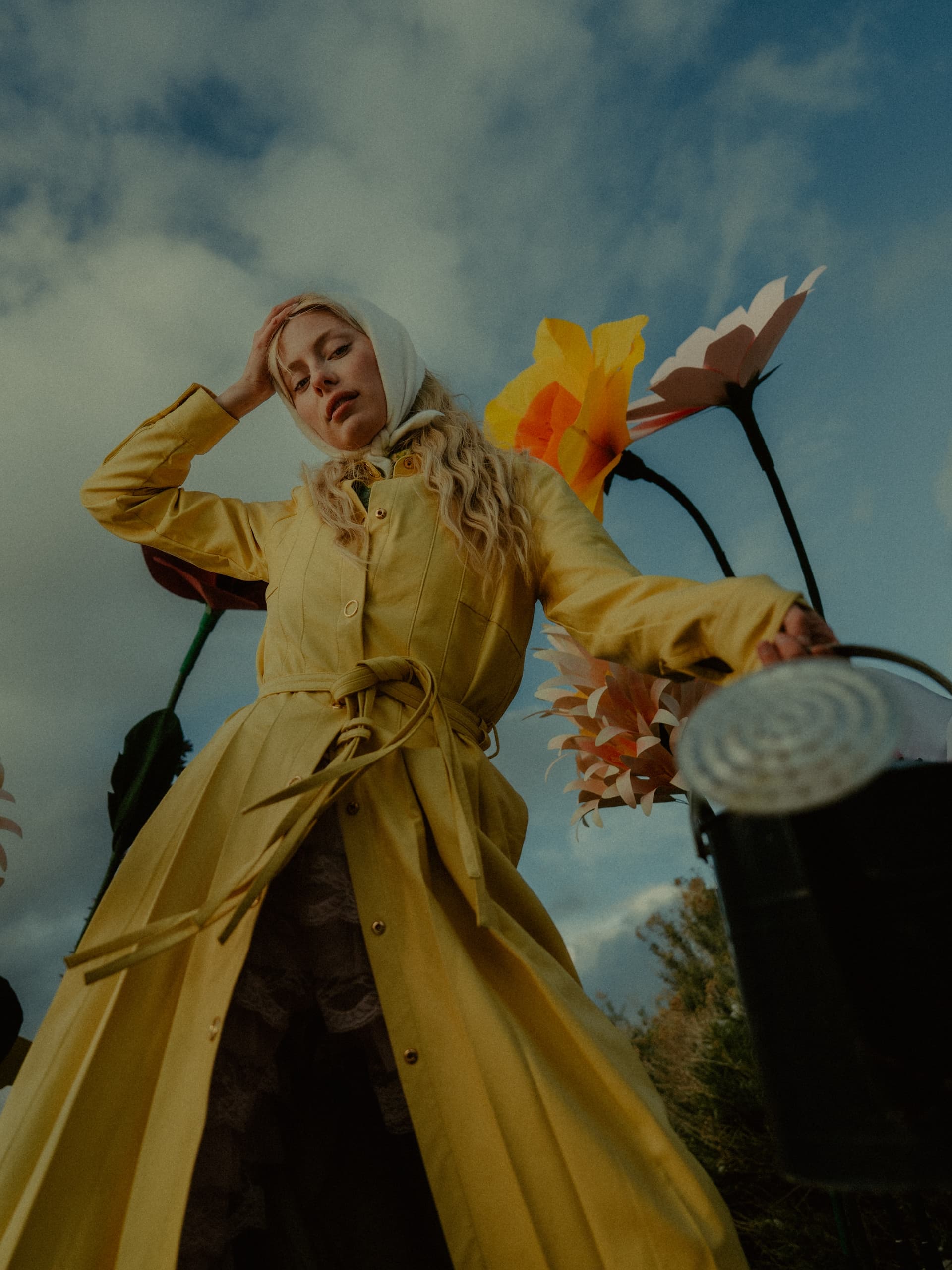 Woman in yellow coat standing with flowers and watering can under cloudy sky.