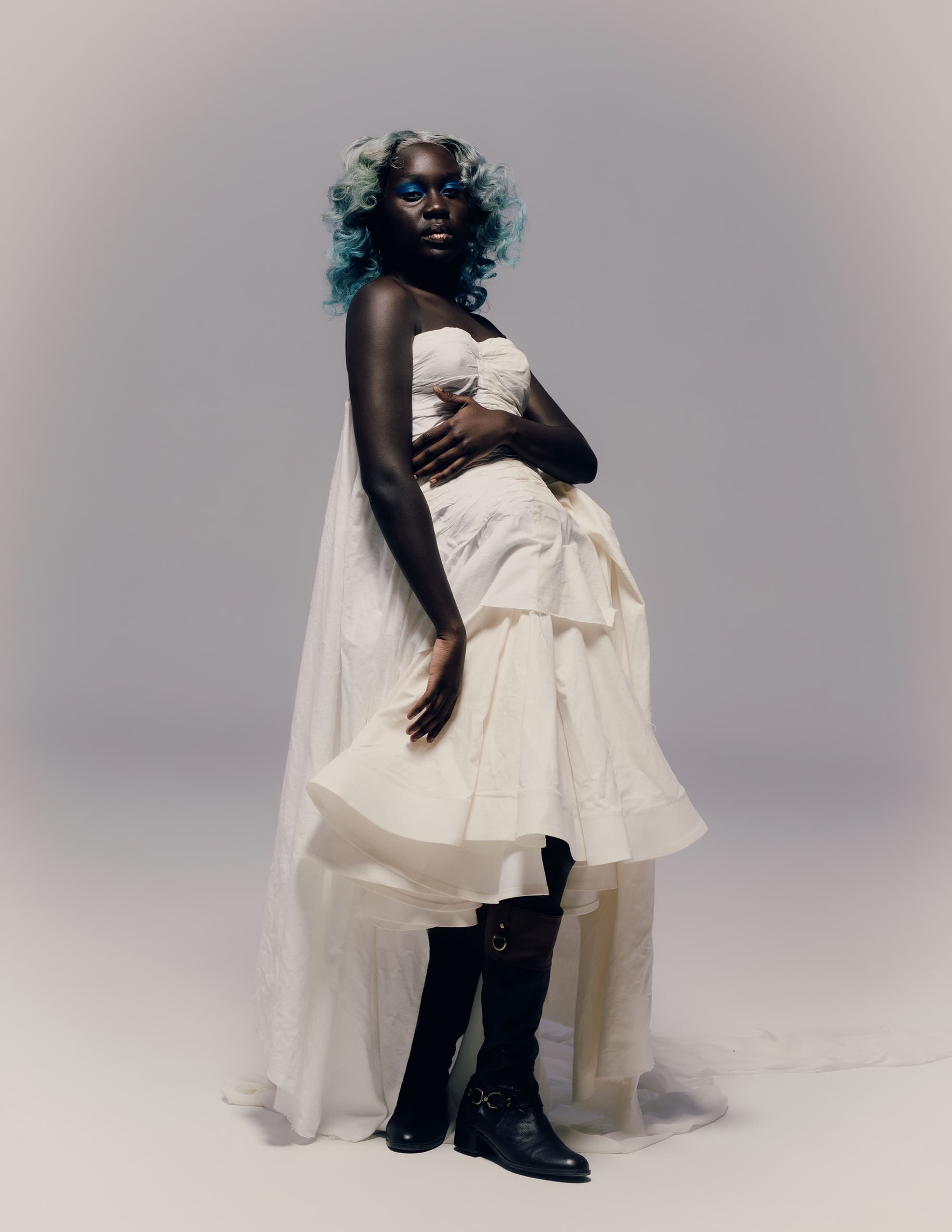 Stylish model in white dress with blue hair and boots, striking a confident pose.