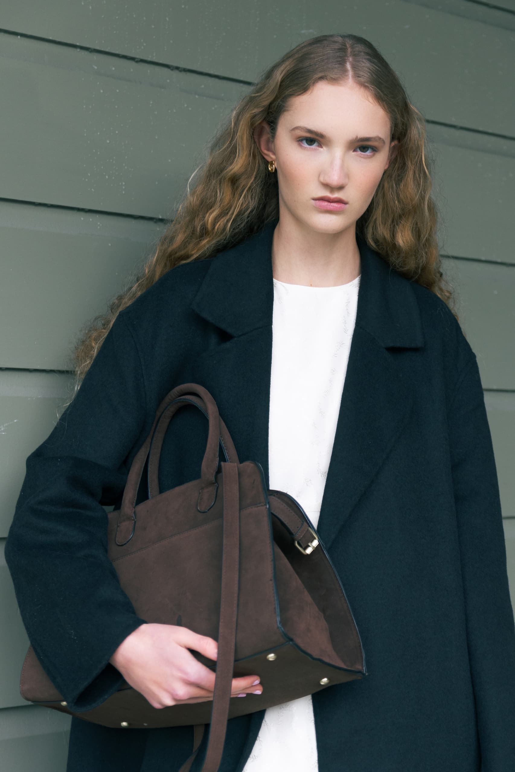Person in elegant coat holding a stylish brown handbag against a green wall.