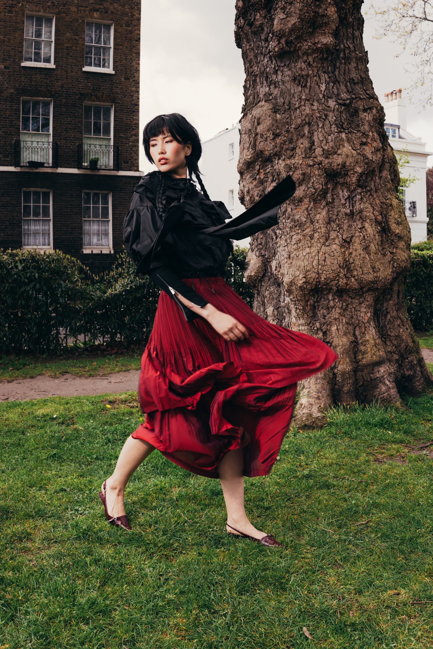 Woman twirling in red skirt near tree in urban park setting.