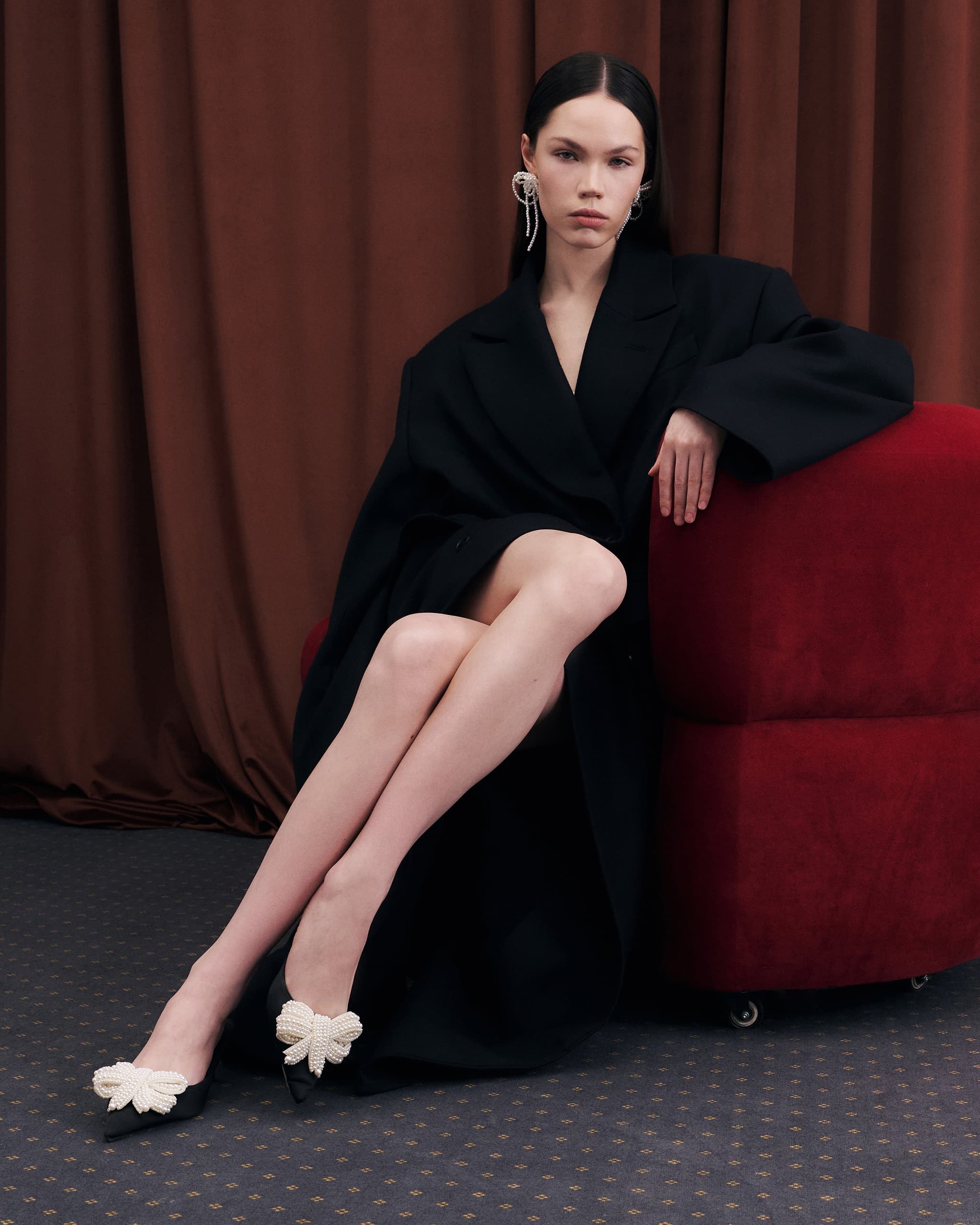 Elegant woman in black coat and pearl-embellished shoes sitting on a red chair.