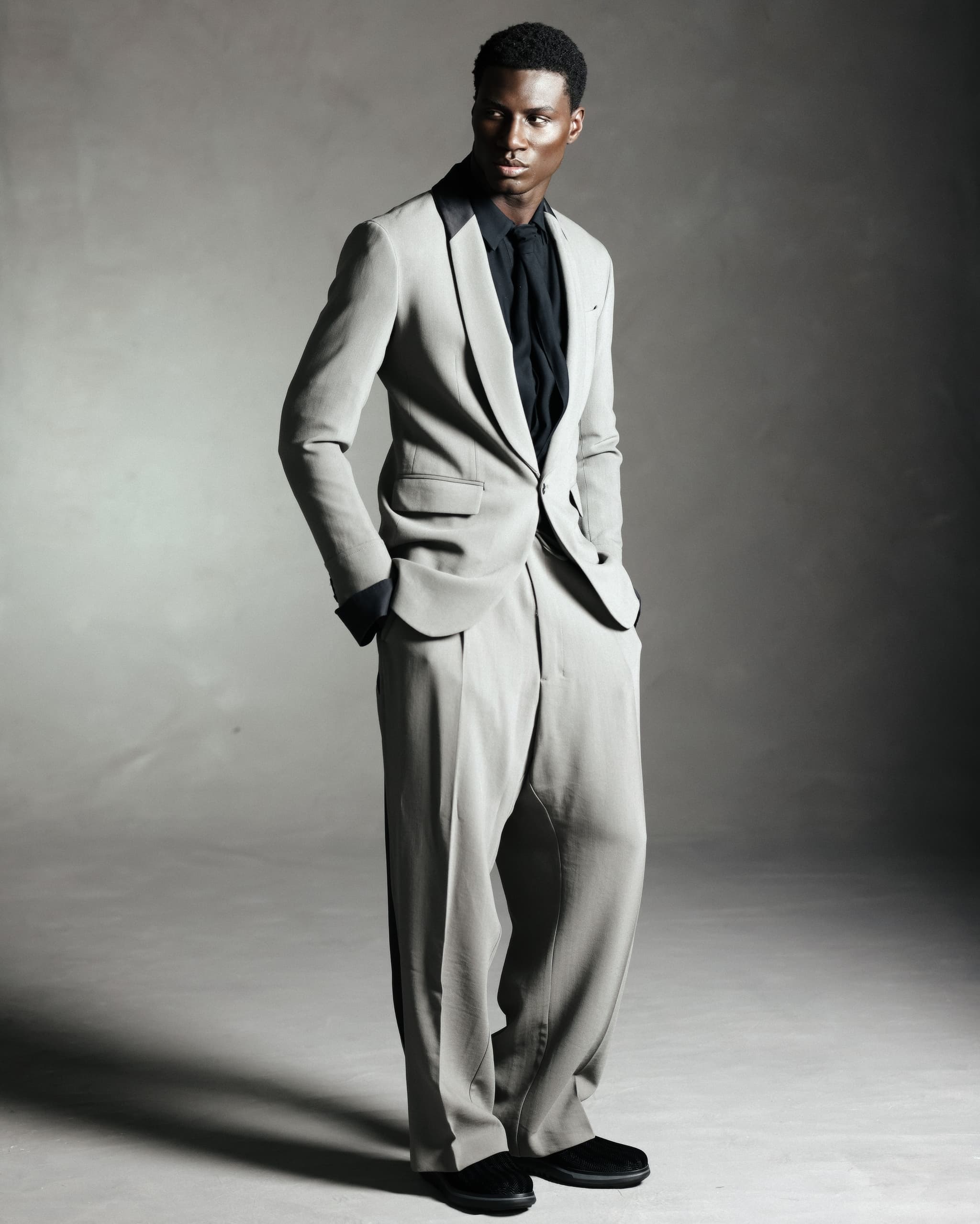 Man in elegant gray suit with black shirt, standing confidently against a neutral backdrop.