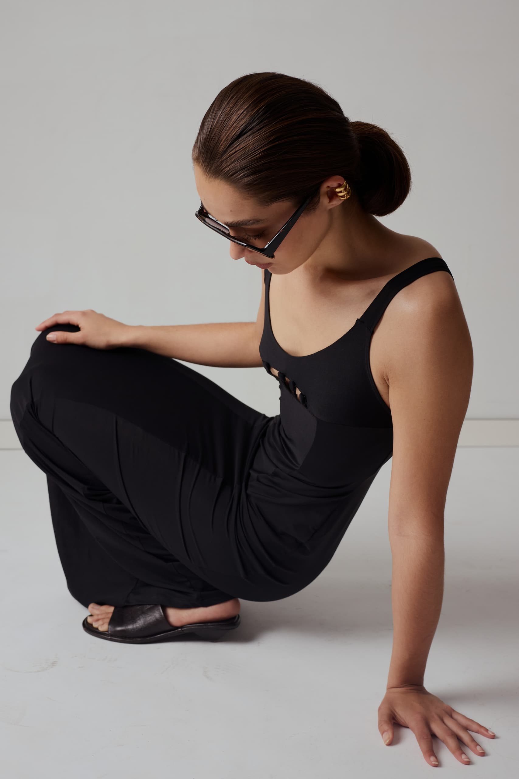 Woman in black dress sits elegantly, wearing sunglasses and gold ear cuff.