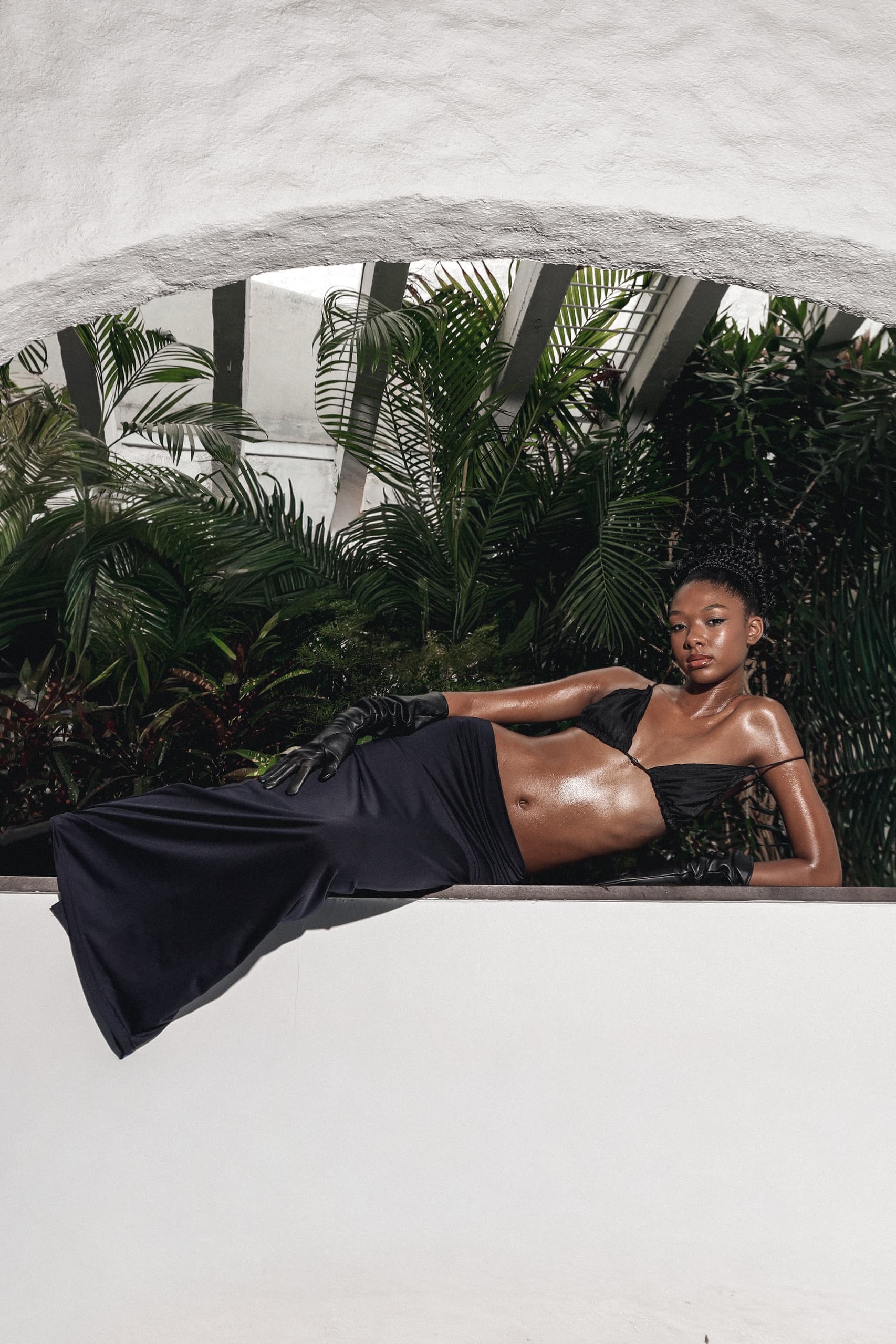 Elegant woman in black dress lounging by lush green foliage.