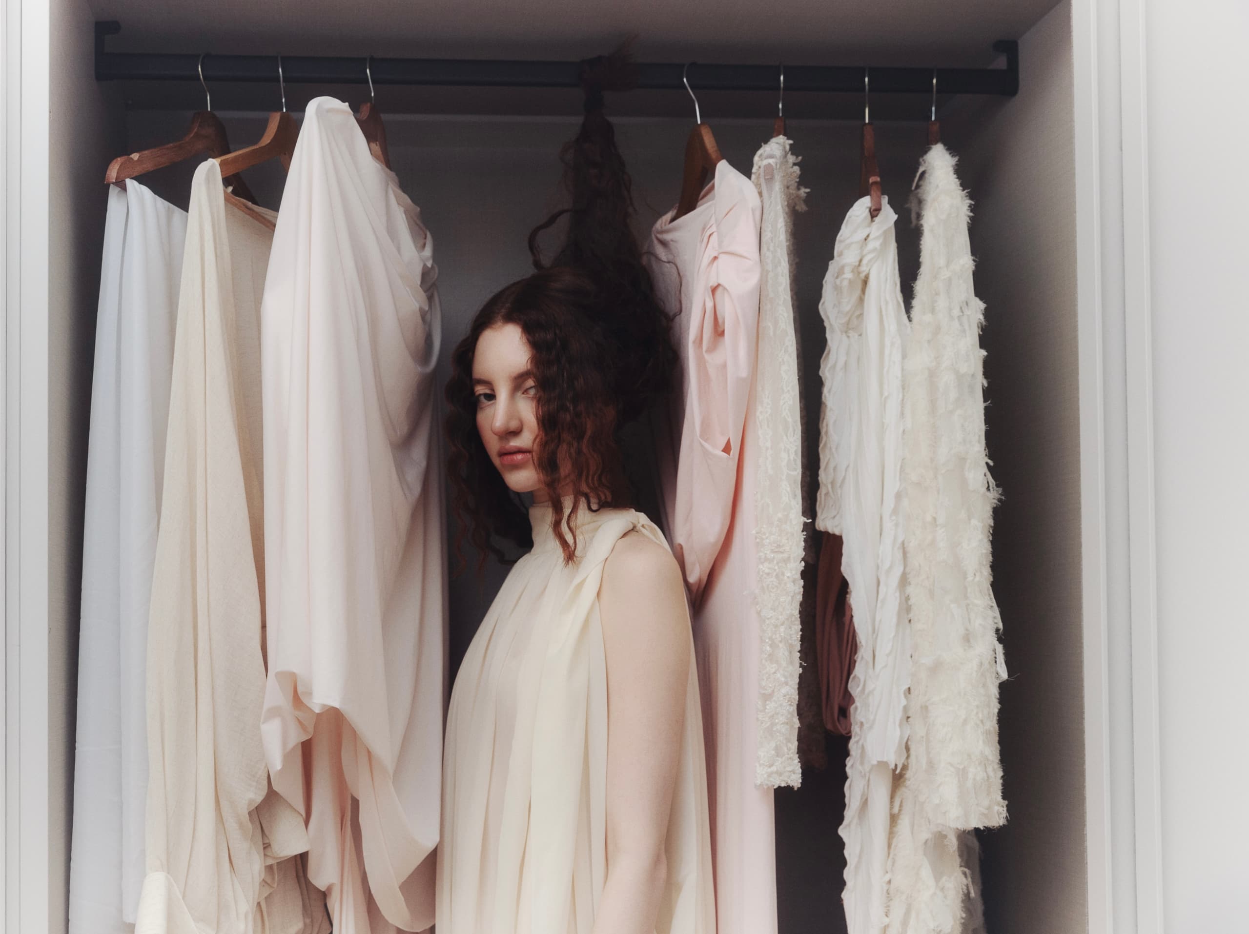 Woman in elegant dress standing in wardrobe among light-colored clothing.