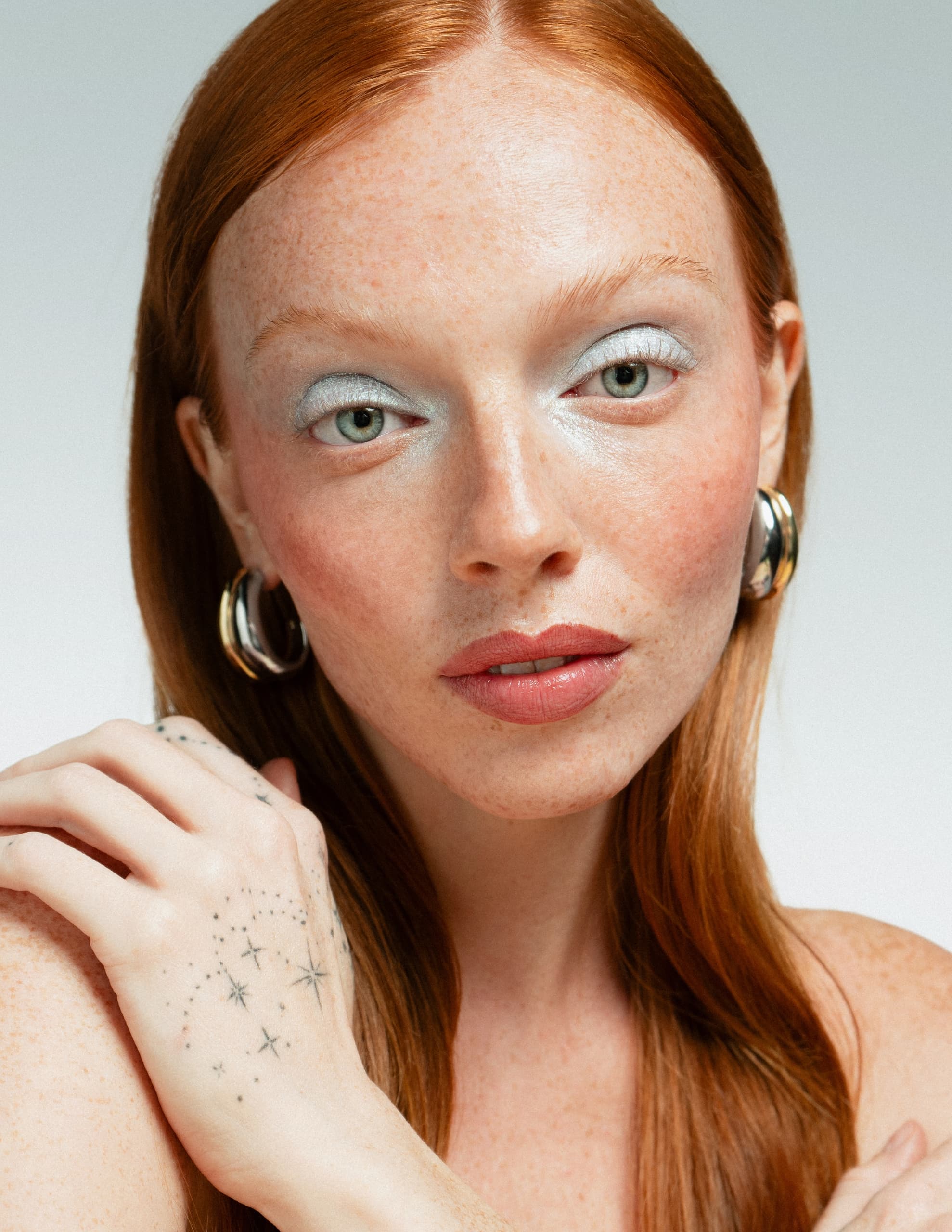 Woman with red hair, freckles, and star hand tattoo, wearing silver hoop earrings.