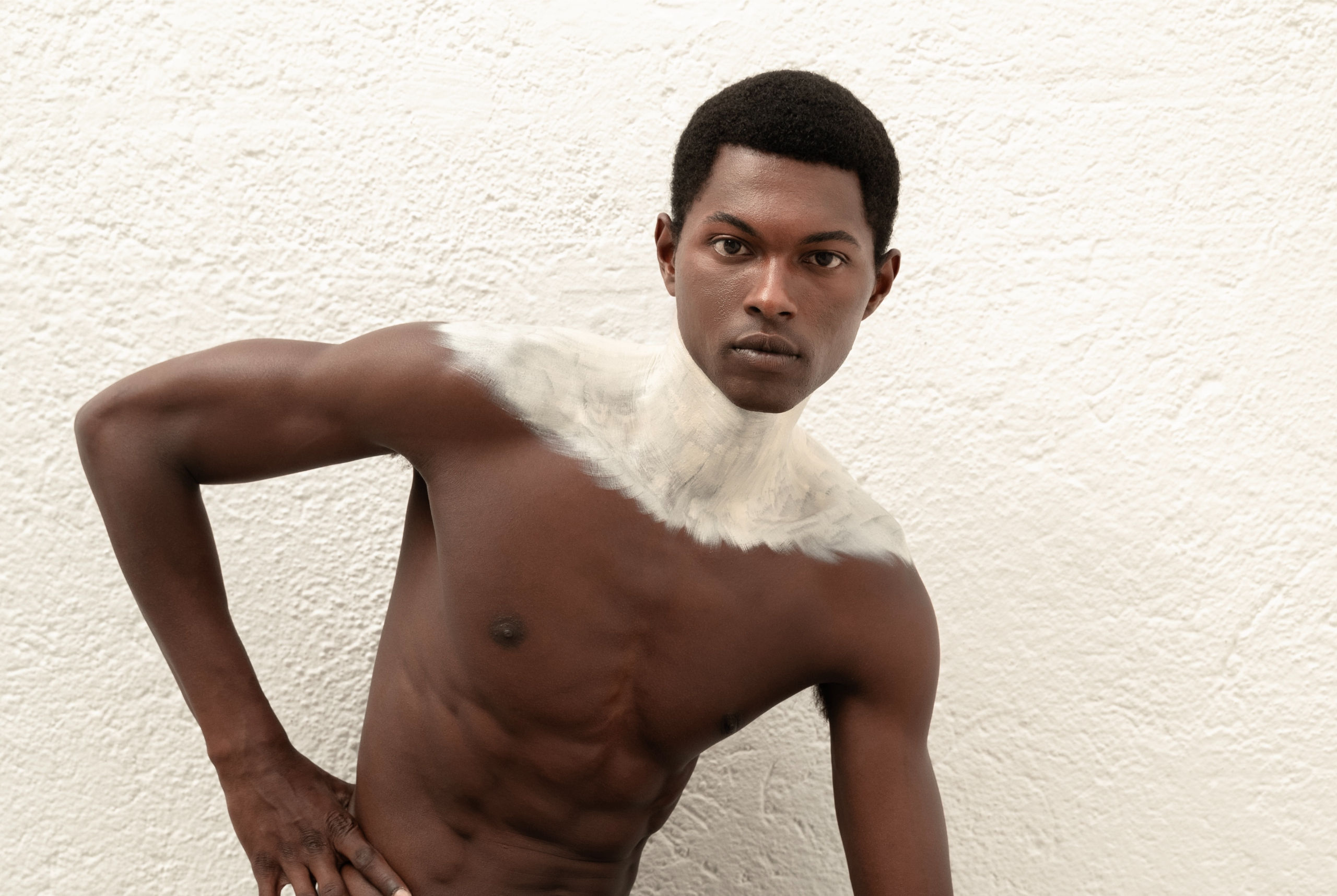 Man with painted shoulders poses against textured white wall.