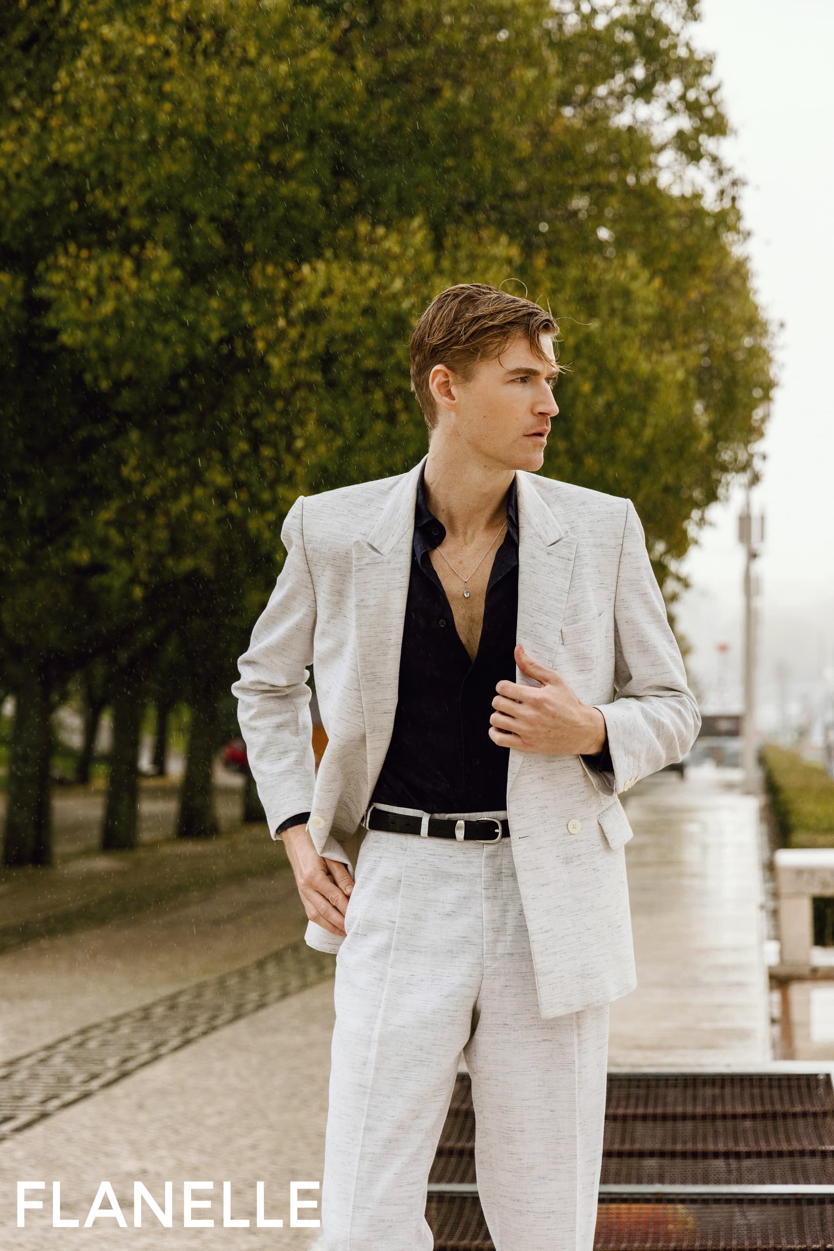 Man in stylish suit walking outdoors, greenery in background.