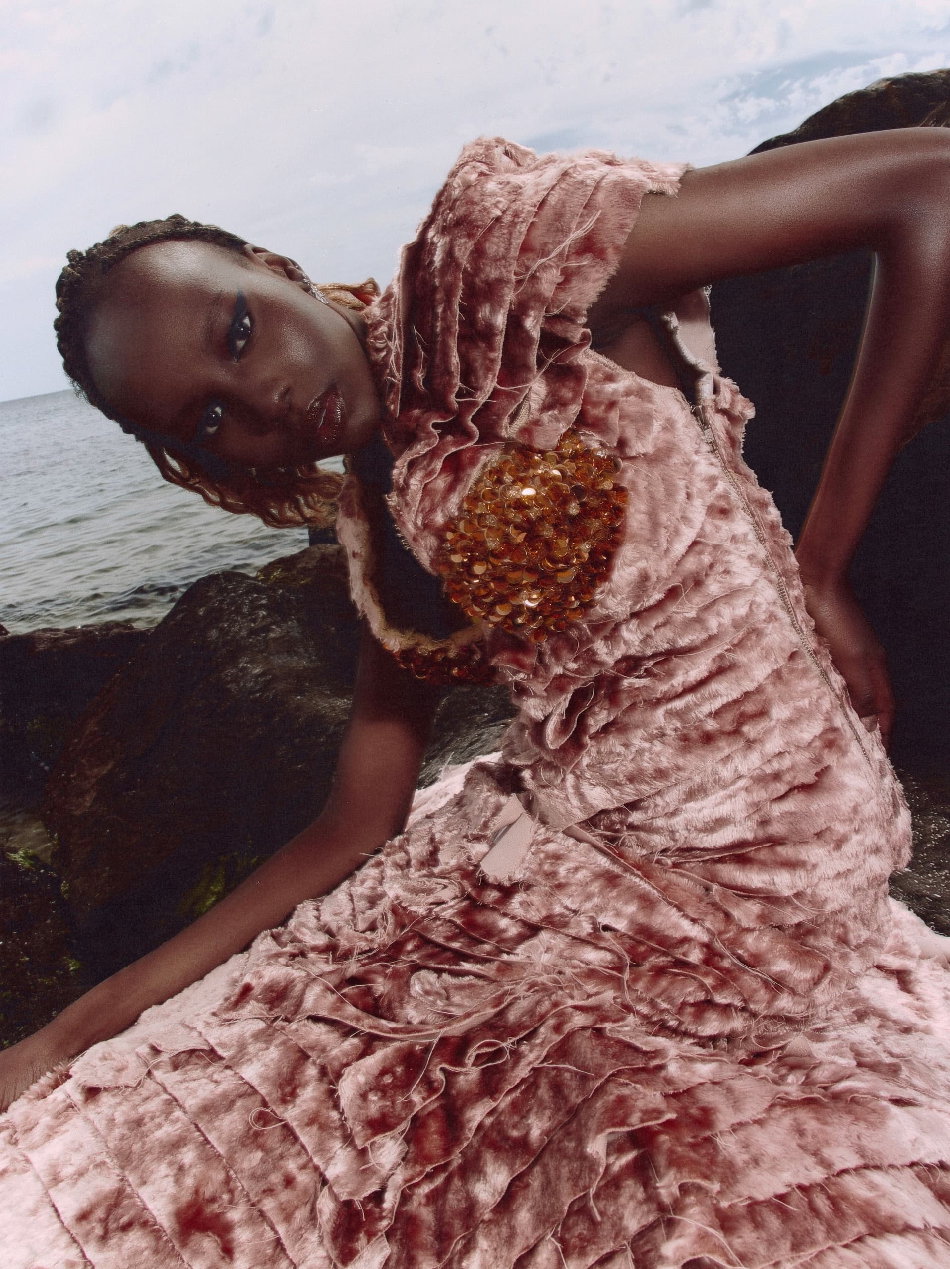 Model in an elegant textured pink dress beside the ocean rocks.