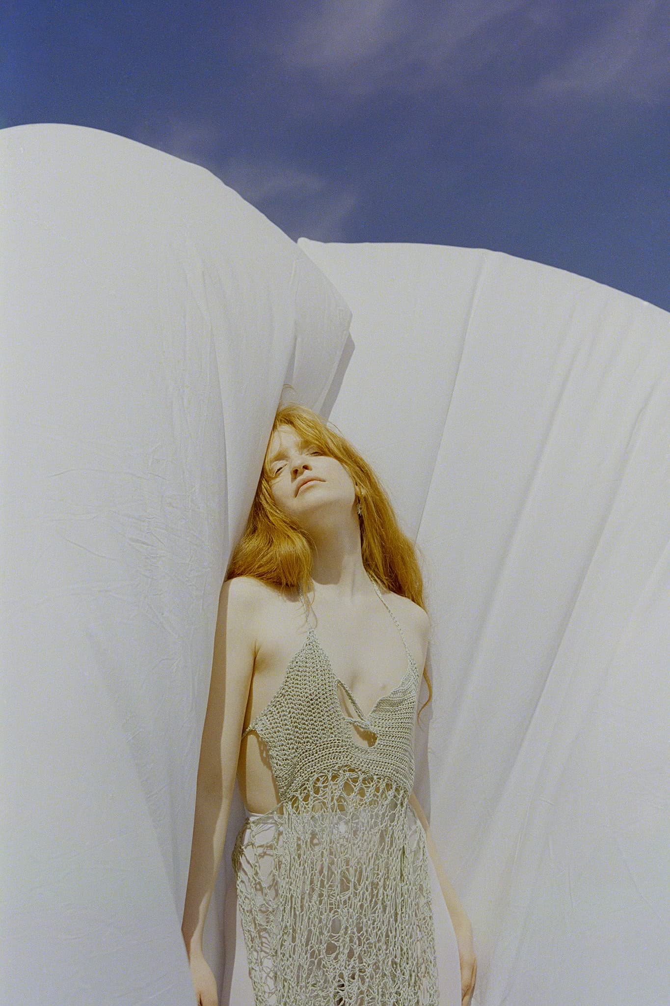 Woman standing outside in crochet dress, leaning on white fabric, under blue sky.